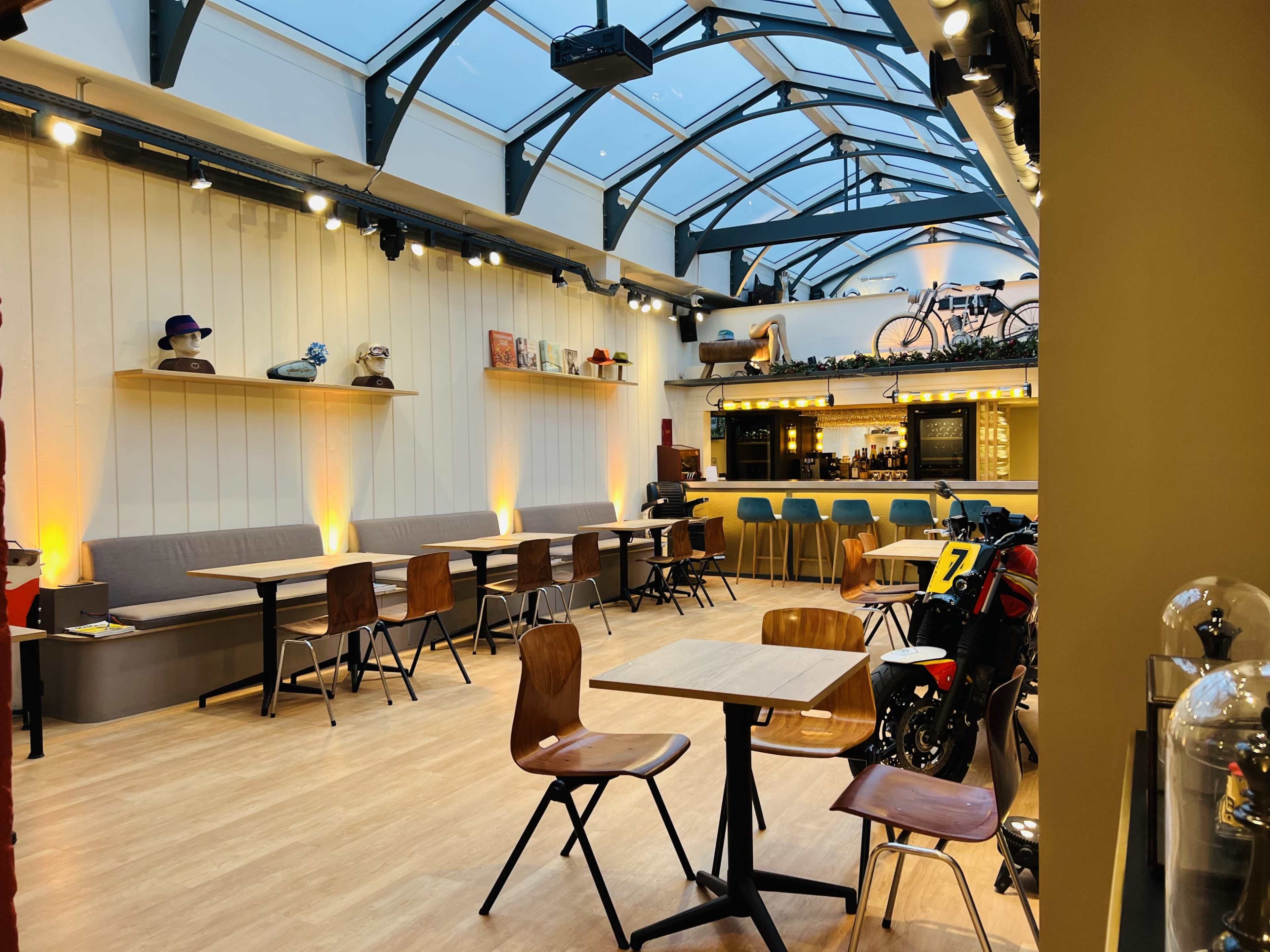 The image shows a brightly lit café with wooden tables and chairs, a bar area in the background, and a glass roof allowing natural light to enter.