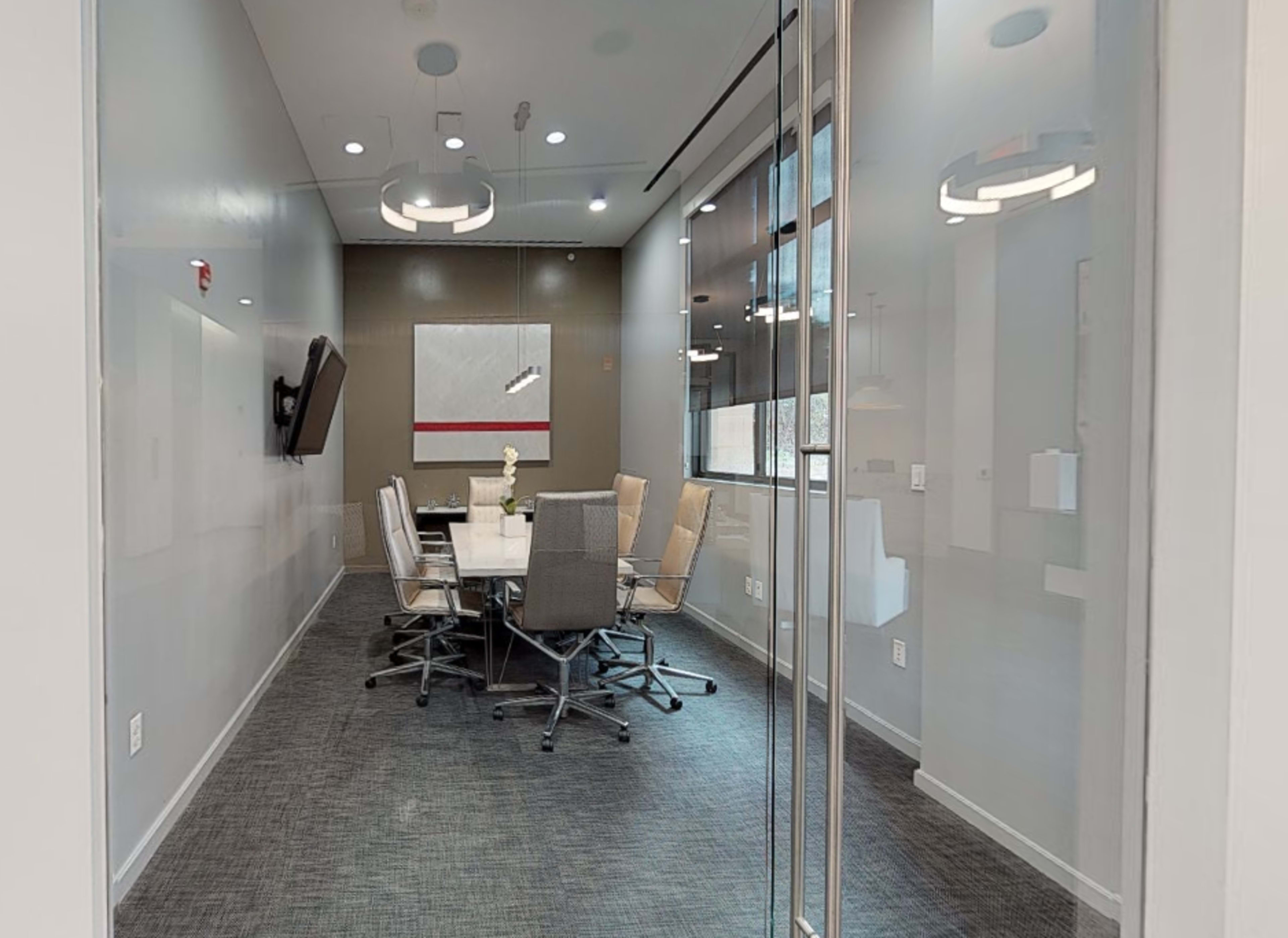 A conference room features a large table surrounded by six chairs, with a television mounted on the wall and large windows allowing natural light.