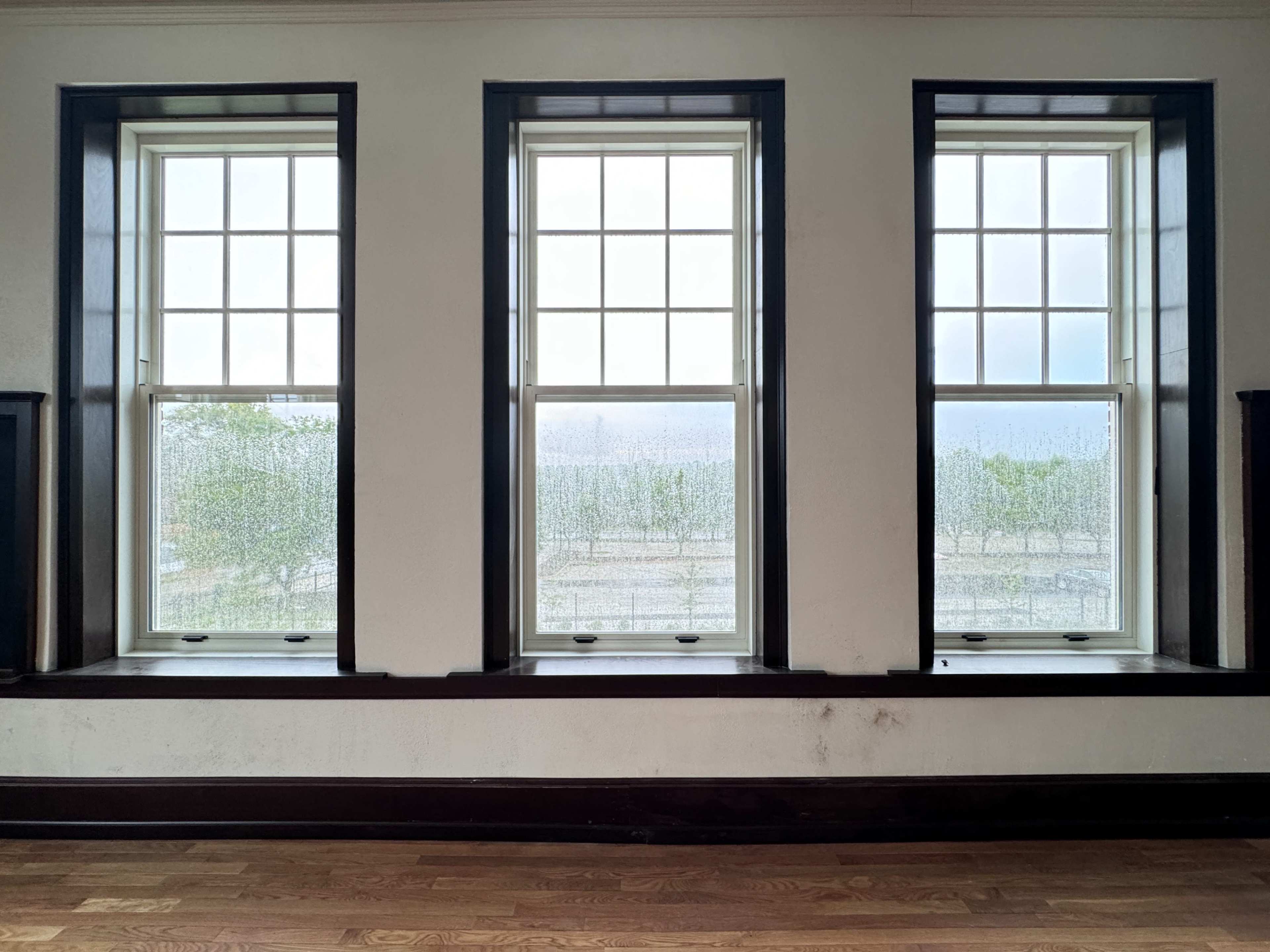 Three windows with rain-streaked glass are set in a wall, revealing a distant view through a cloudy haze.