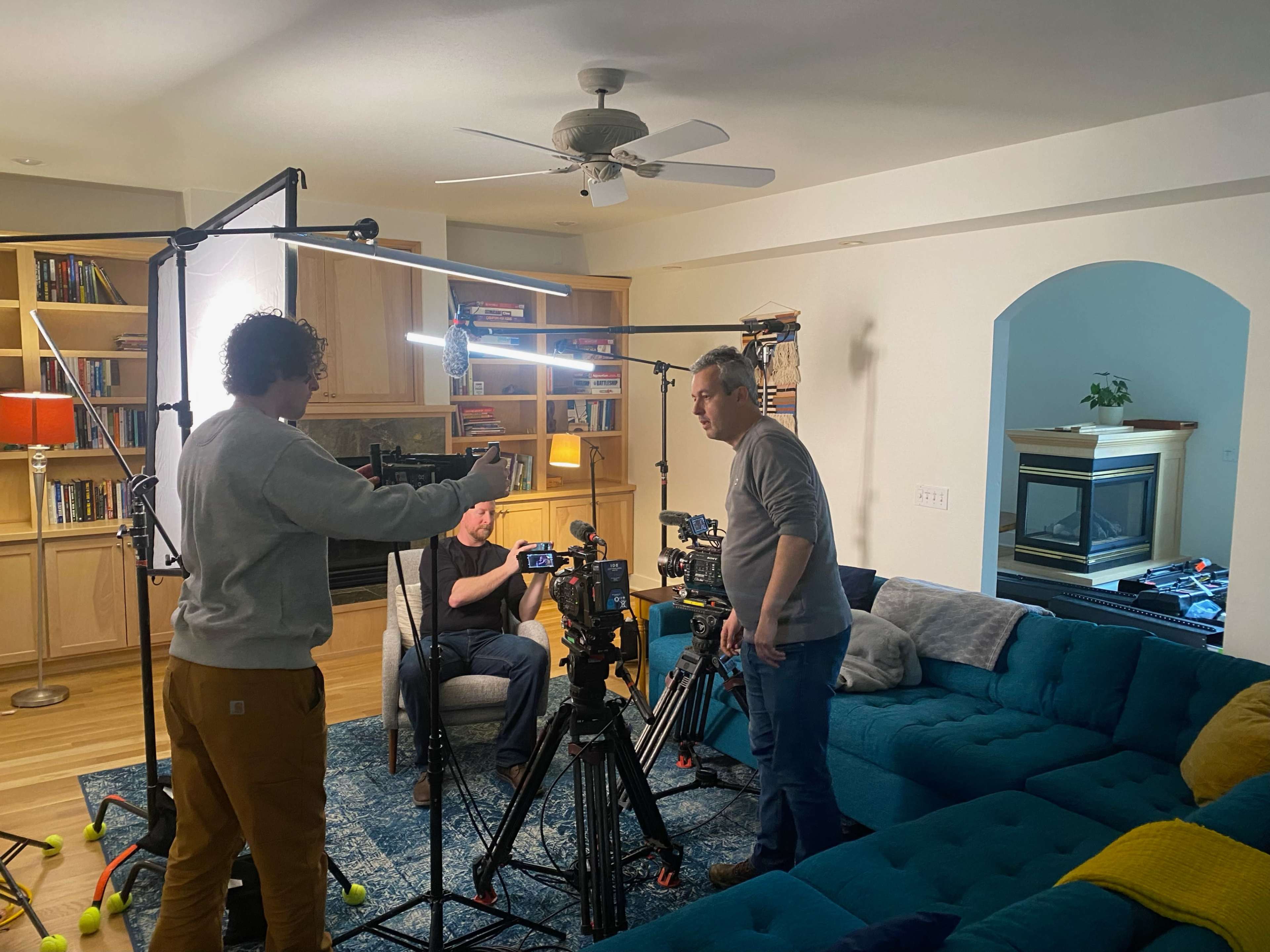 Three men set up filming equipment in a living room with bookshelves and a blue couch.