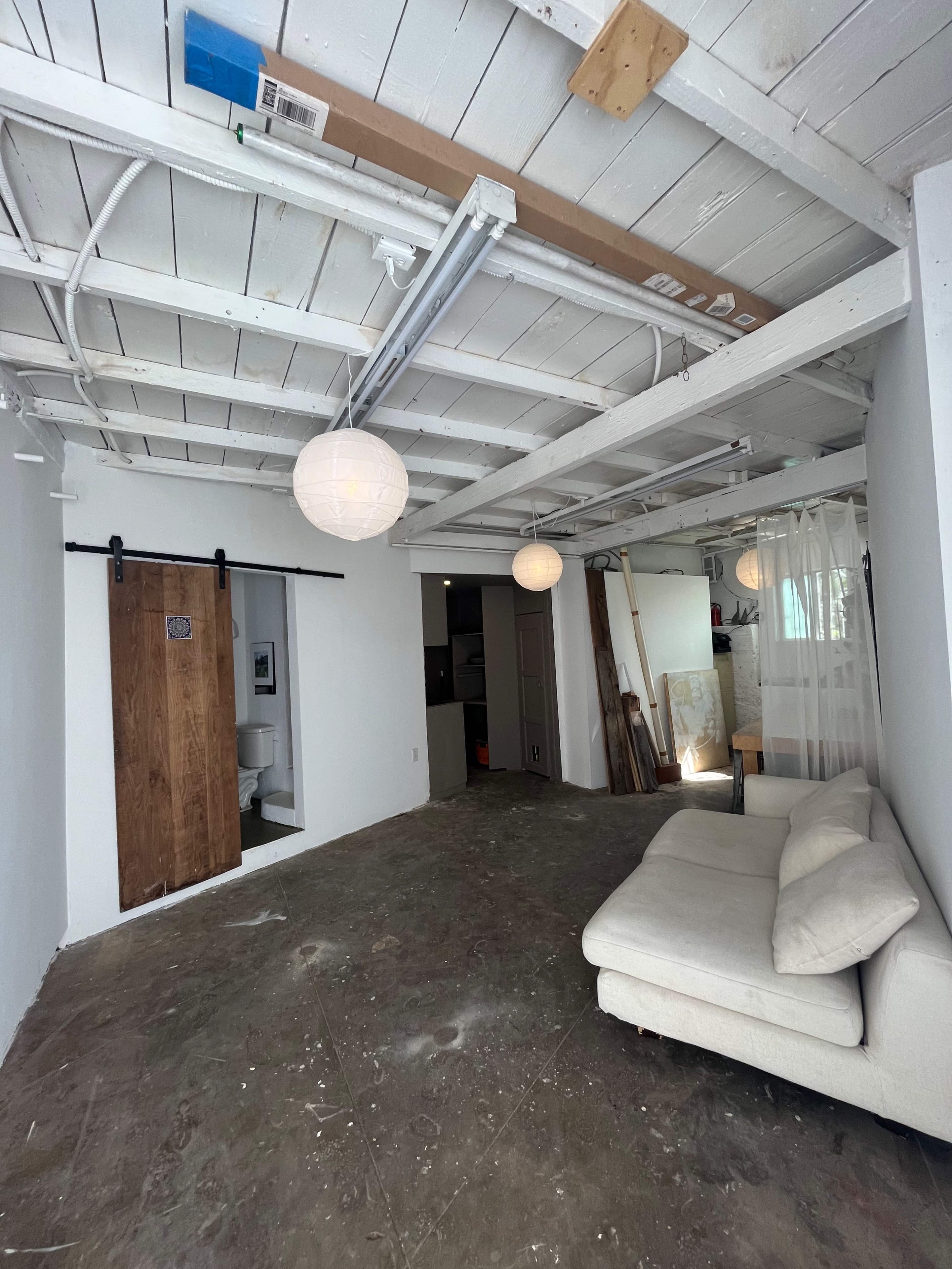 A minimalist interior space features a white sofa, exposed beams, hanging light fixtures, and a wooden sliding door leading to a bathroom.