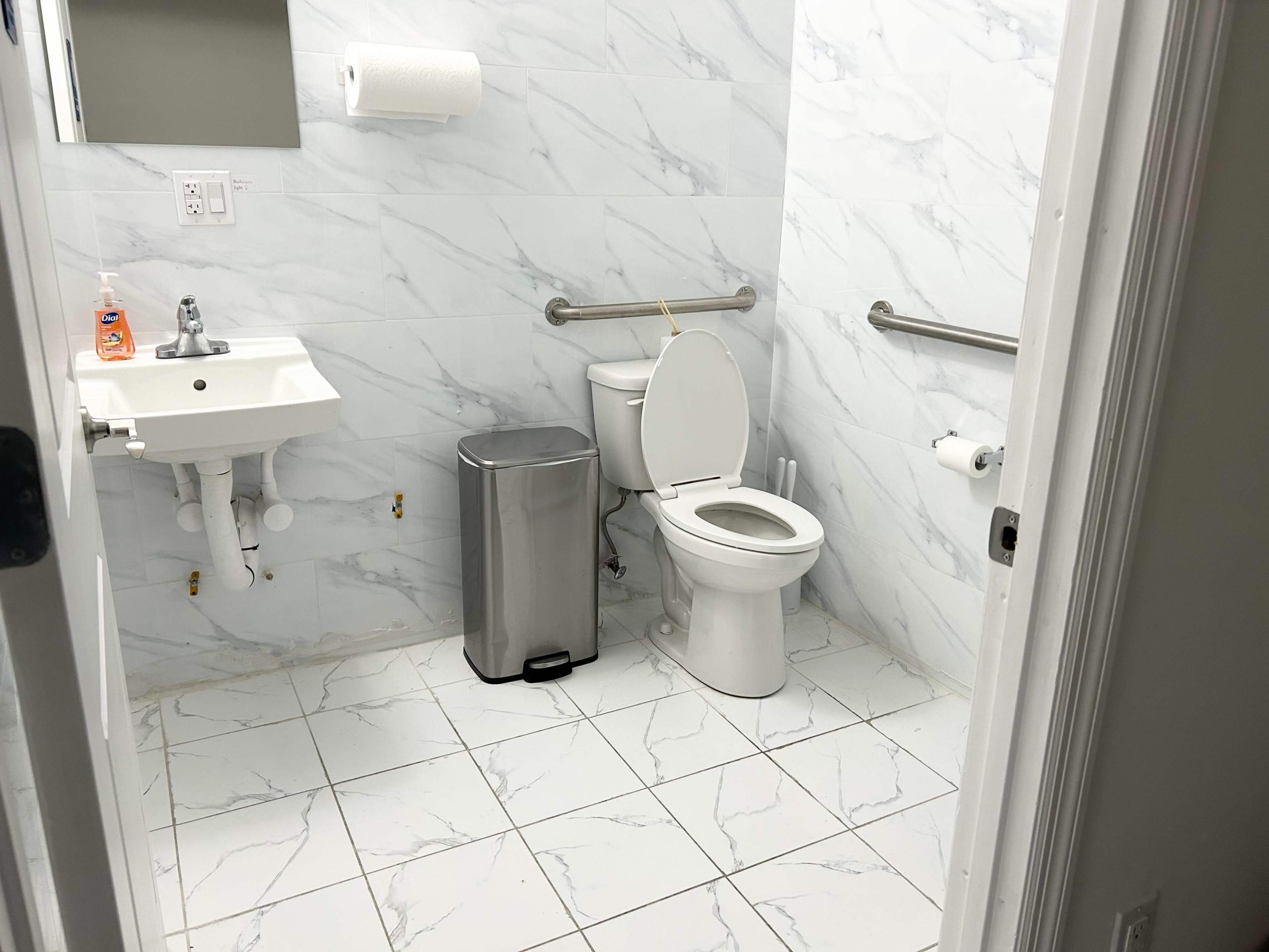 A small, clean bathroom featuring a toilet, a sink, a trash can, and grab bars mounted on the walls.