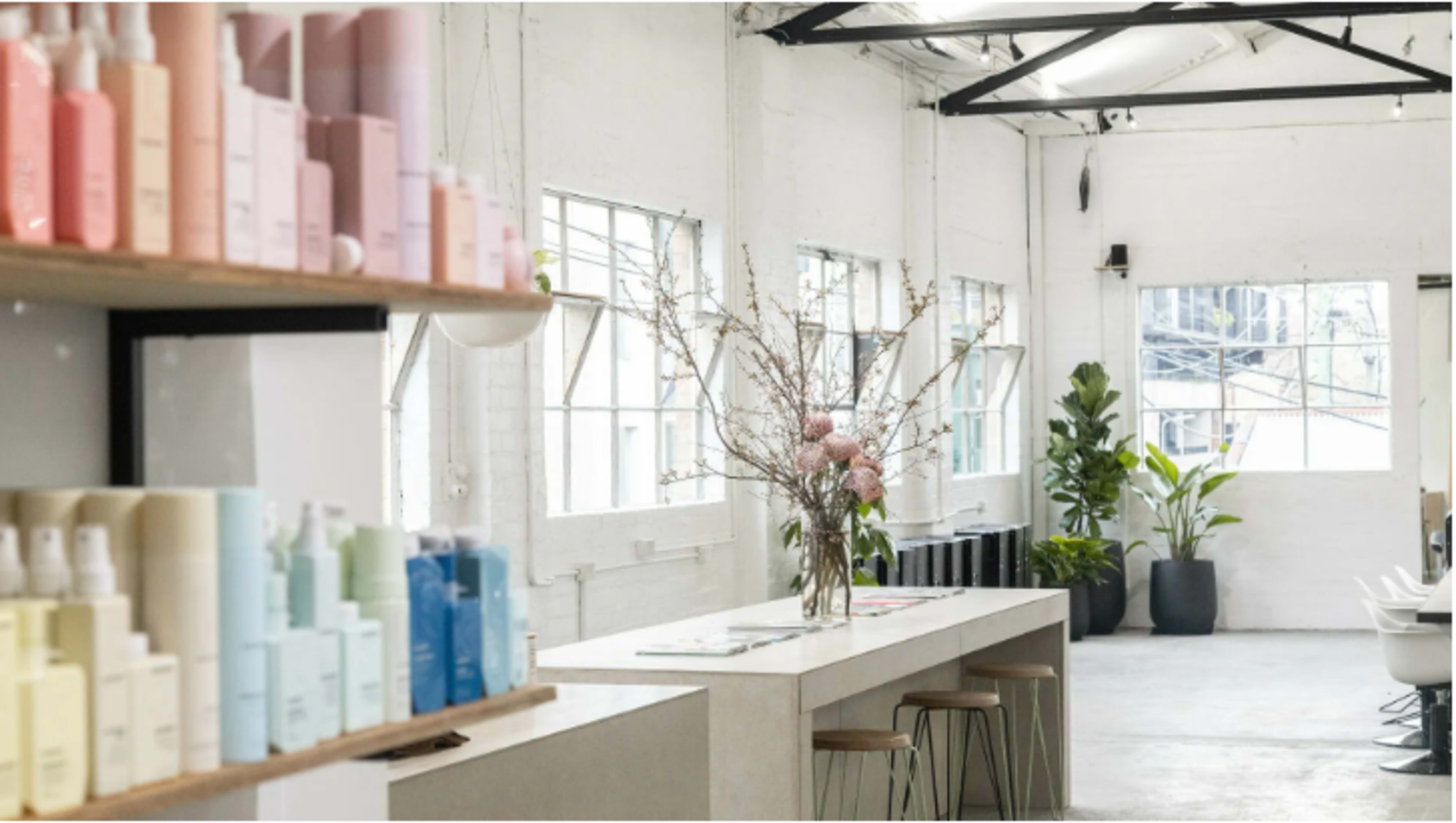 The image shows a bright, modern interior of a beauty or skincare store with shelves displaying pastel-colored products and a central counter topped with a floral arrangement.