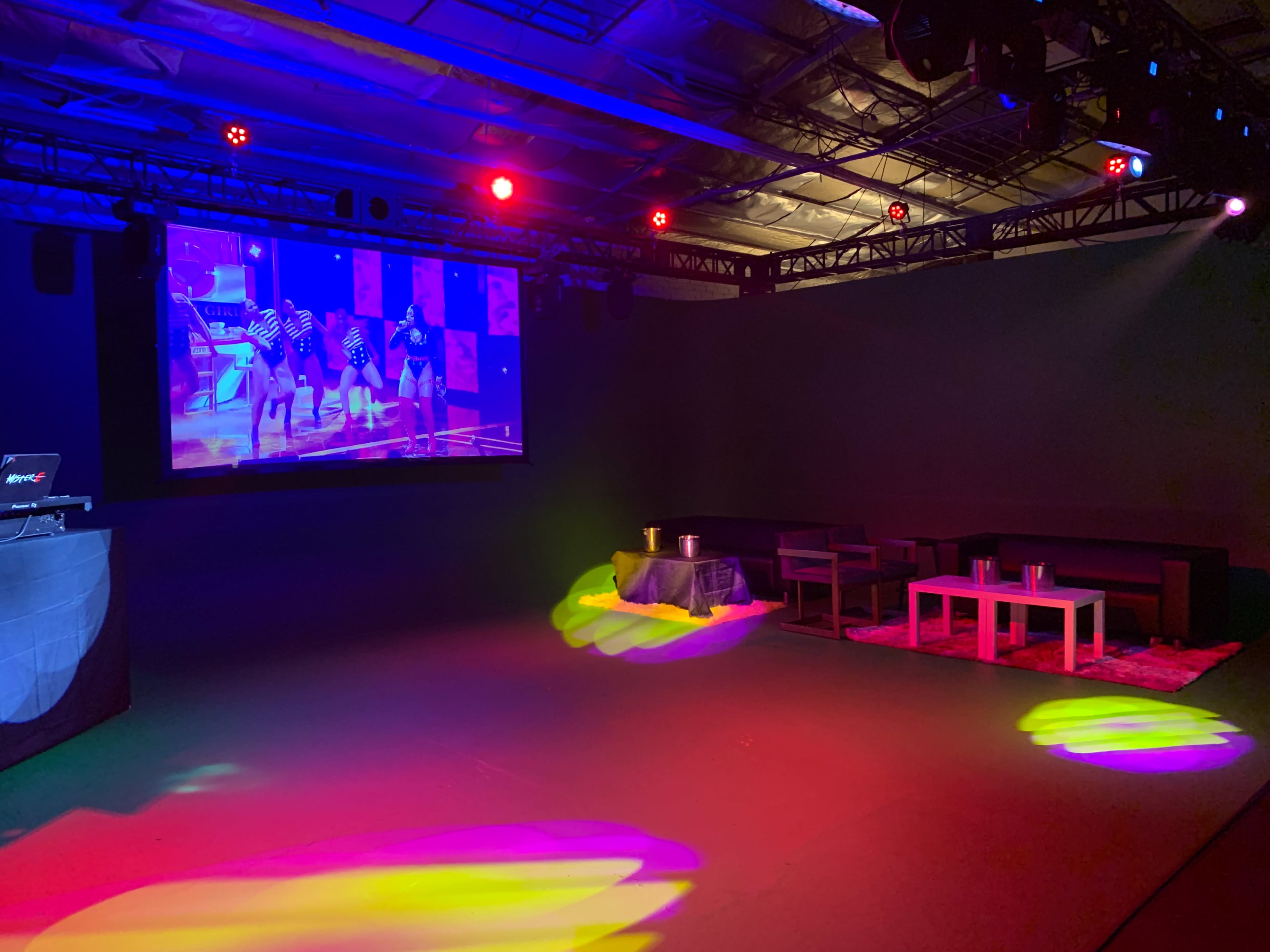 A dimly lit event space features a large screen displaying dancers, with colorful lights illuminating the floor and furniture.