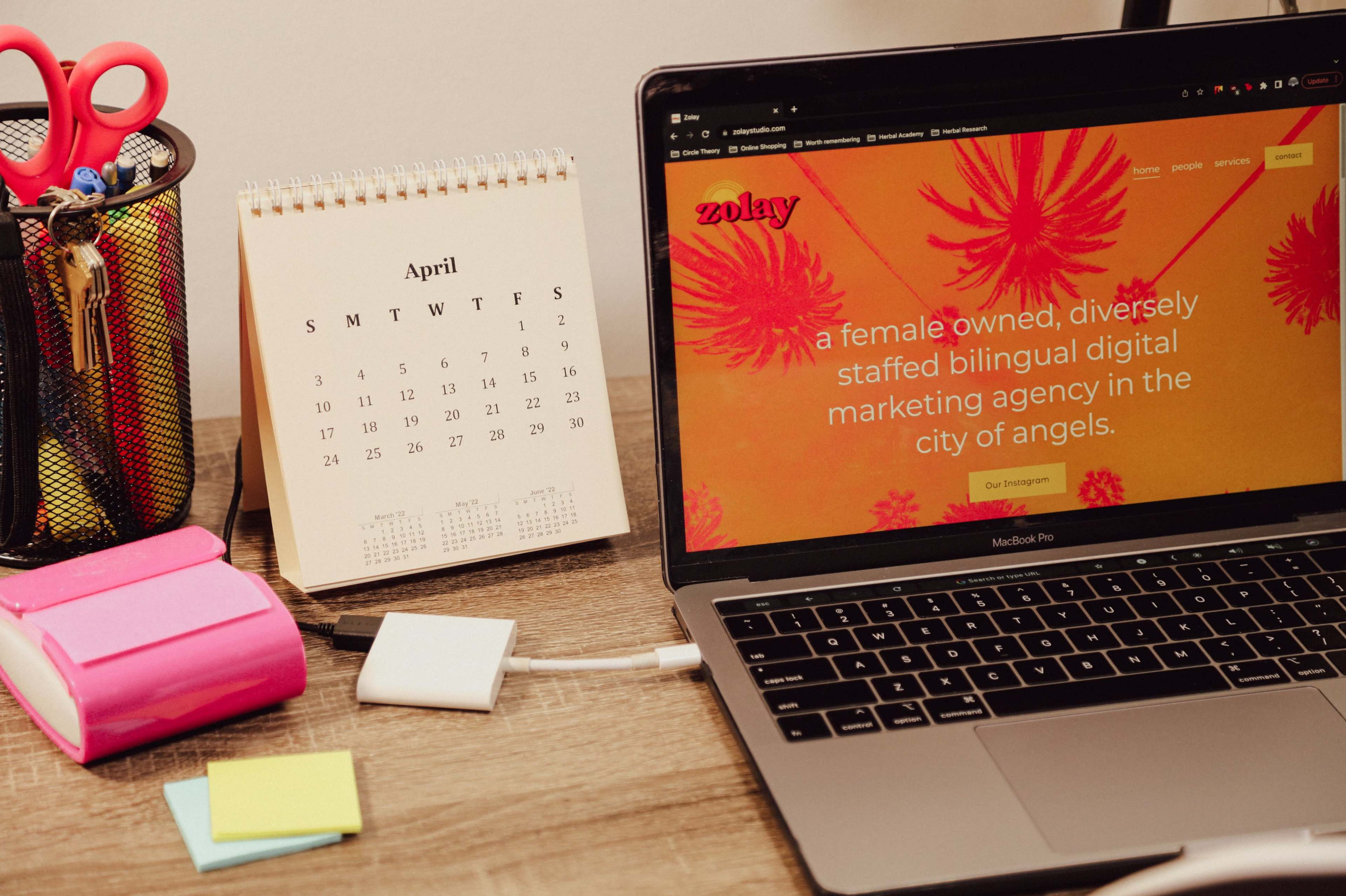 A desk setup features a calendar displaying April, a pink notepad, a charging device, and a laptop open to a marketing agency's website with a vibrant orange background.