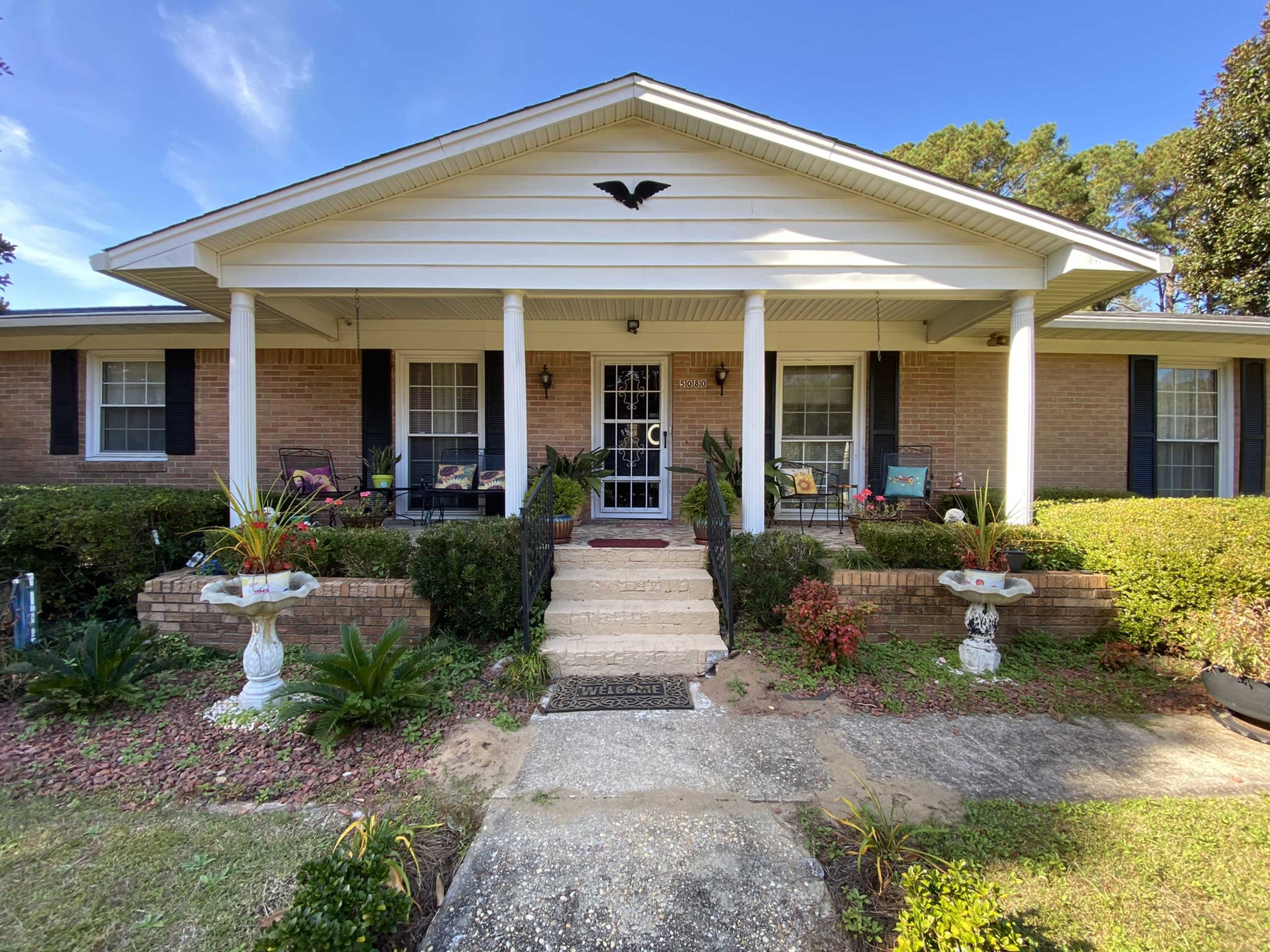 A single-story brick house with a front porch, white columns, and well-maintained landscaping.
