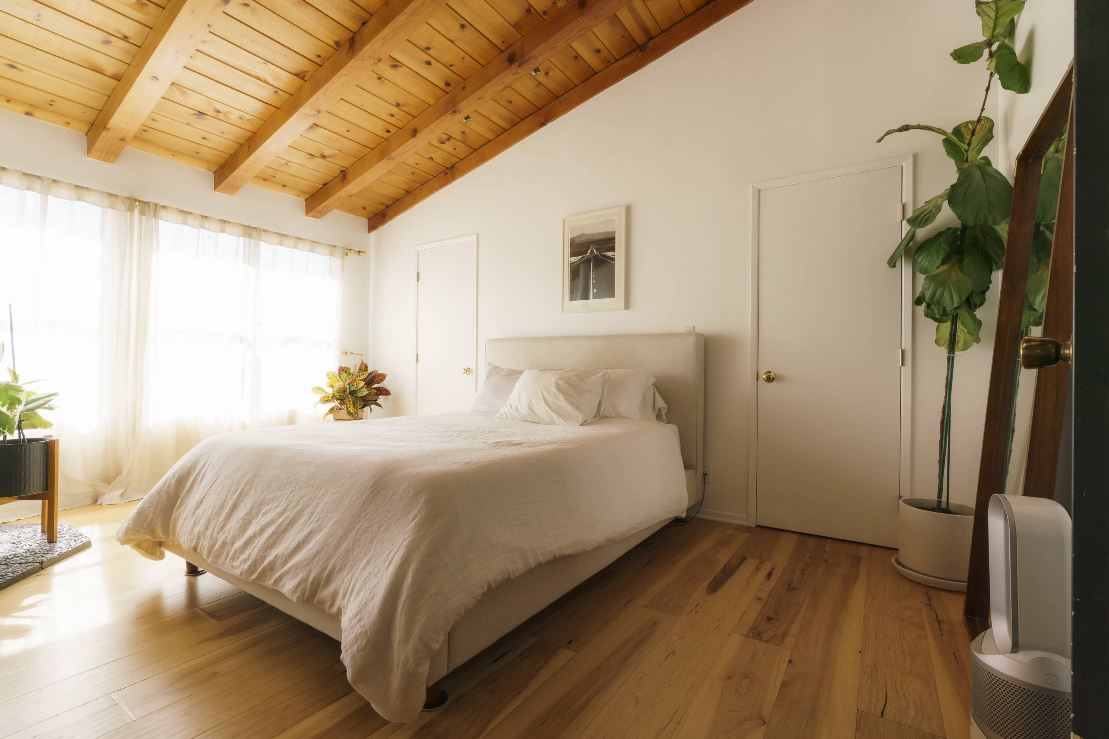 A neatly arranged bedroom features a bed against a wall with wooden beams overhead and large windows allowing natural light to fill the space.
