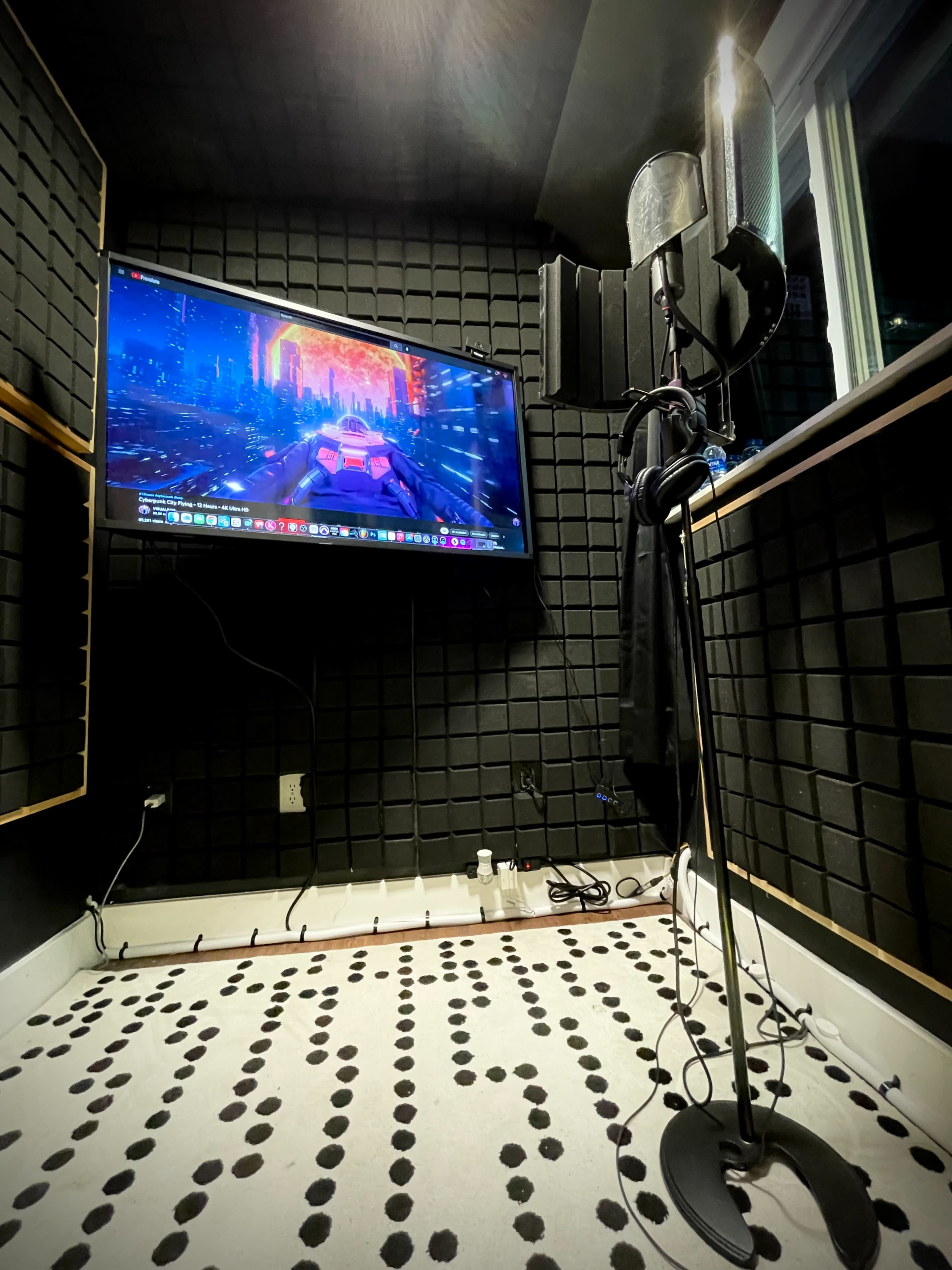 A recording studio features a microphone setup in front of a wall-mounted monitor displaying vibrant graphics, surrounded by soundproofing panels.