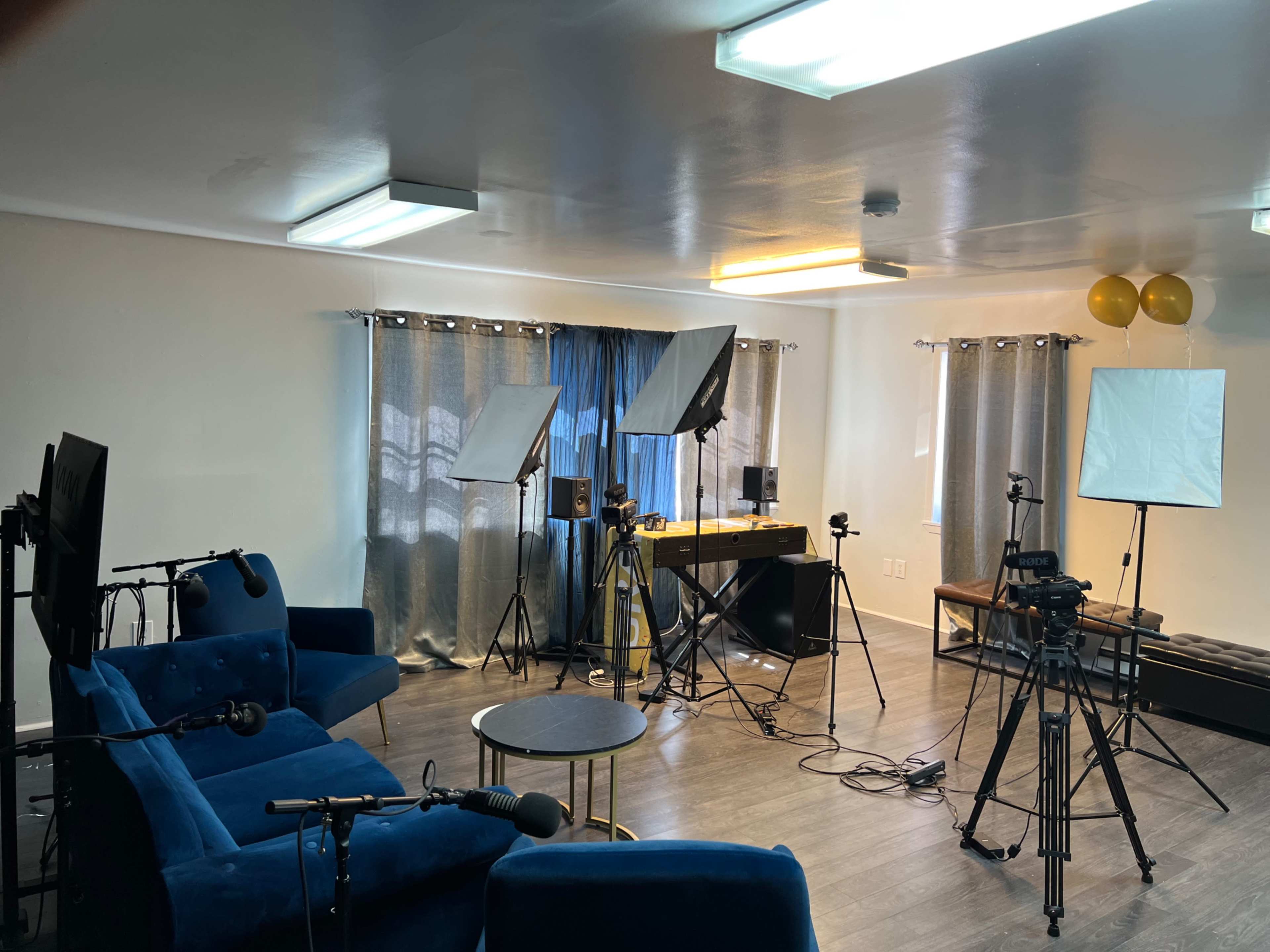 The room is set up for filming, with blue furniture, lighting equipment, and multiple cameras positioned around a table.