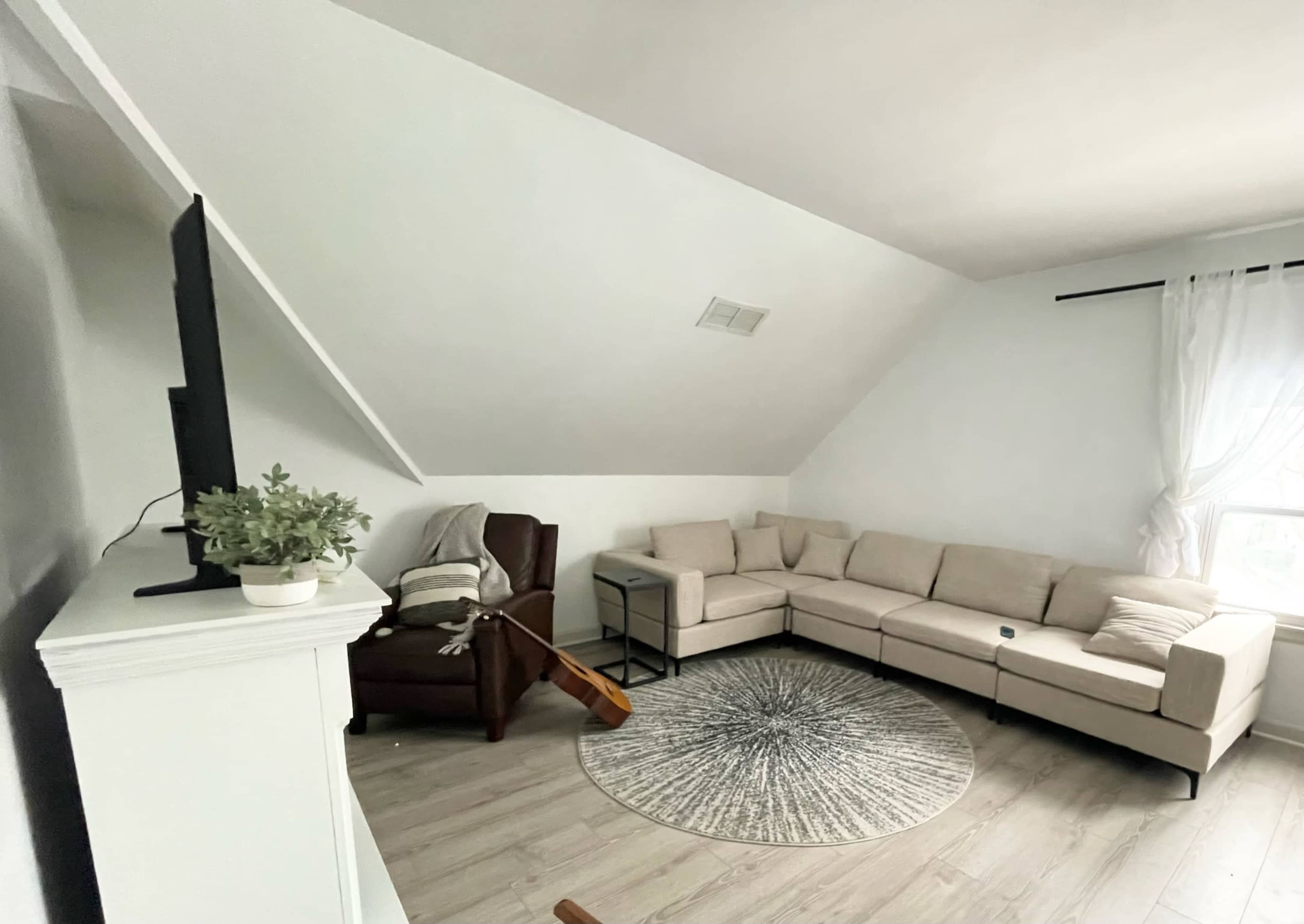 A light-filled living room features a beige sectional sofa, a brown armchair, and a television mounted on the wall.
