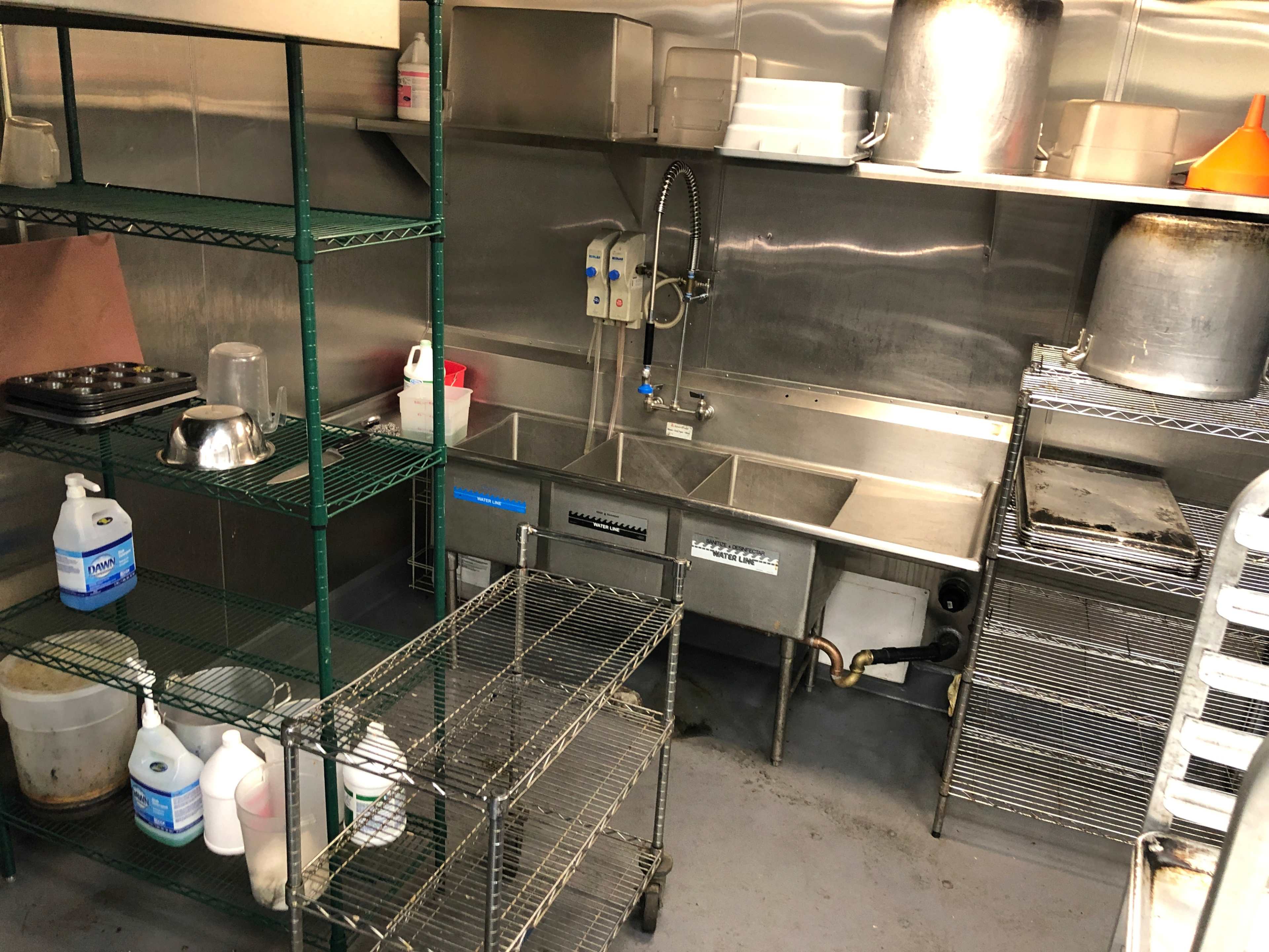 The image shows a commercial kitchen space featuring stainless steel surfaces, shelves with cleaning supplies, and a three-compartment sink.