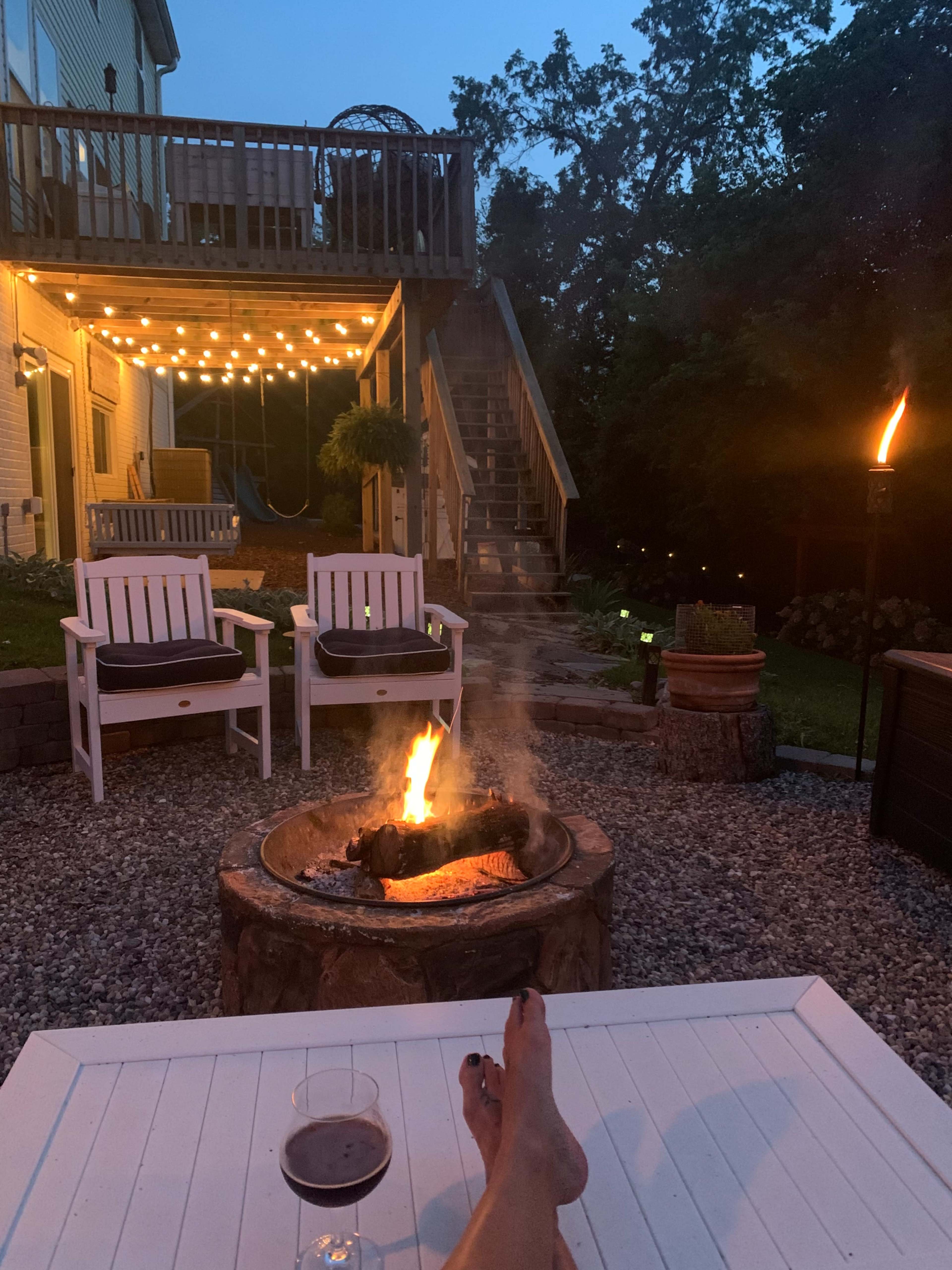 A fire crackles in a stone fire pit surrounded by white chairs on a gravel patio, with a deck illuminated by string lights visible in the background.