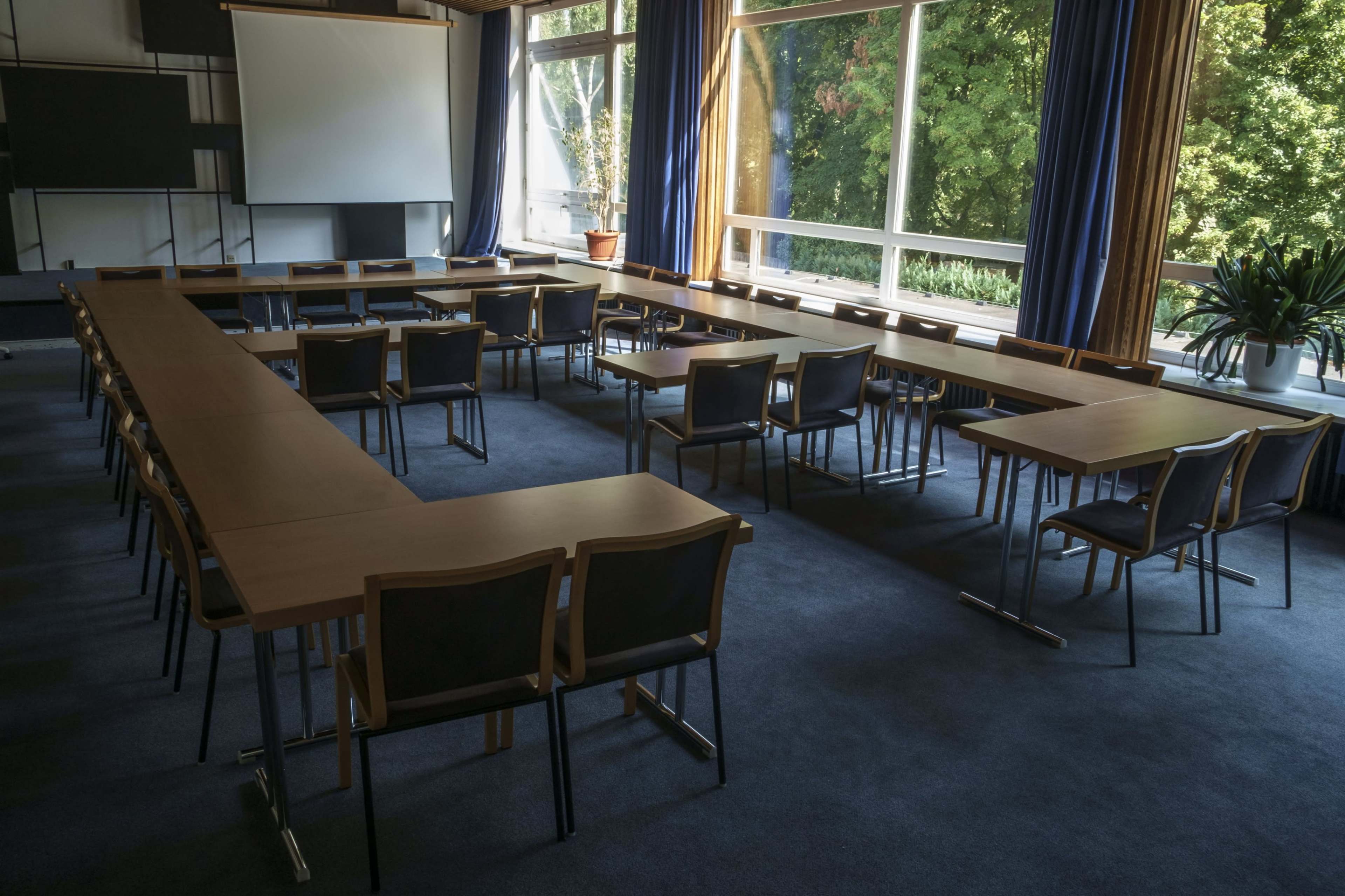 A meeting room is set up with tables arranged in a U-shape and chairs placed around them, featuring large windows that provide natural light and views of greenery outside.