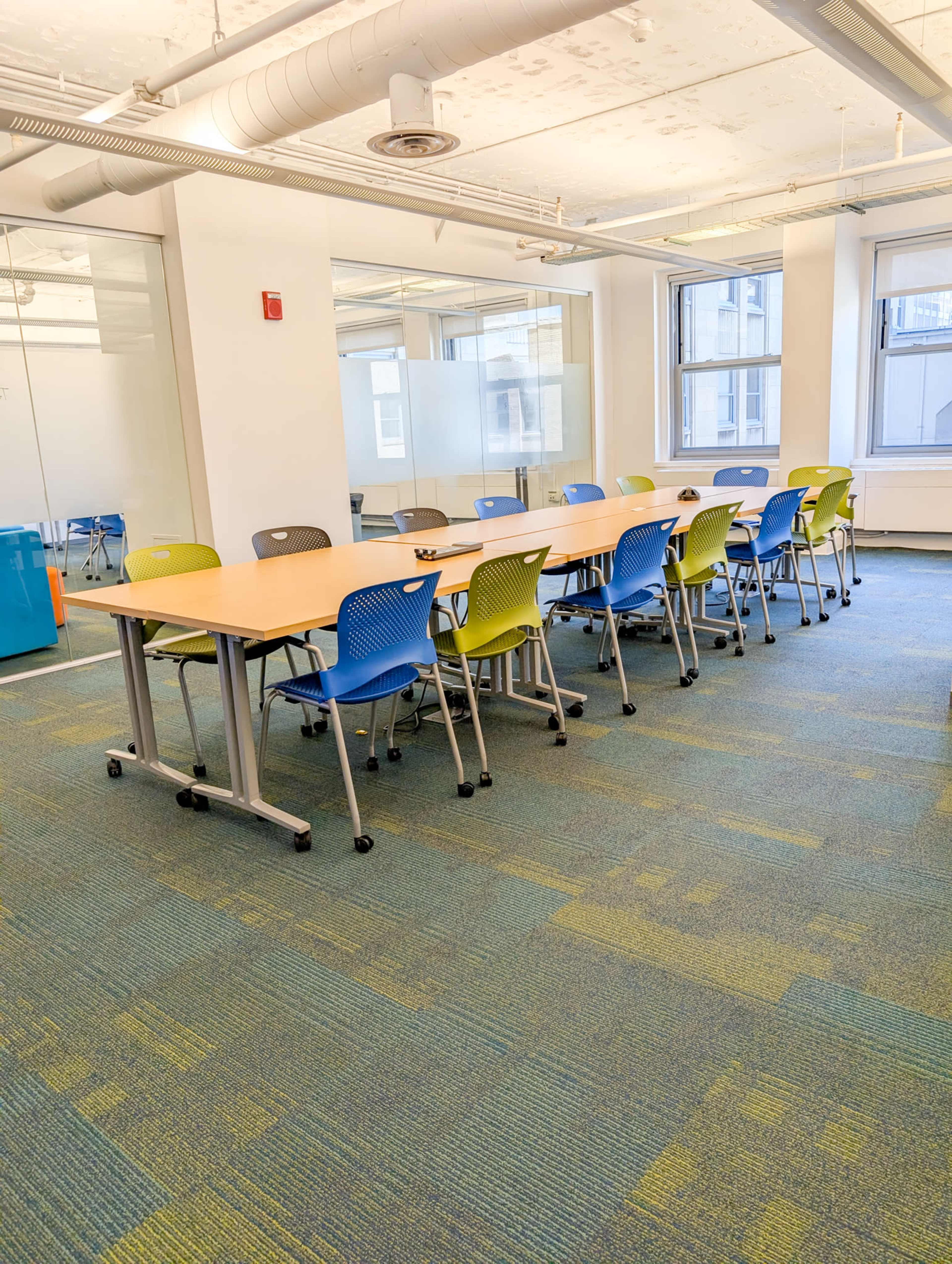 A long table with colorful chairs is positioned in a well-lit office space featuring large windows and a carpeted floor.