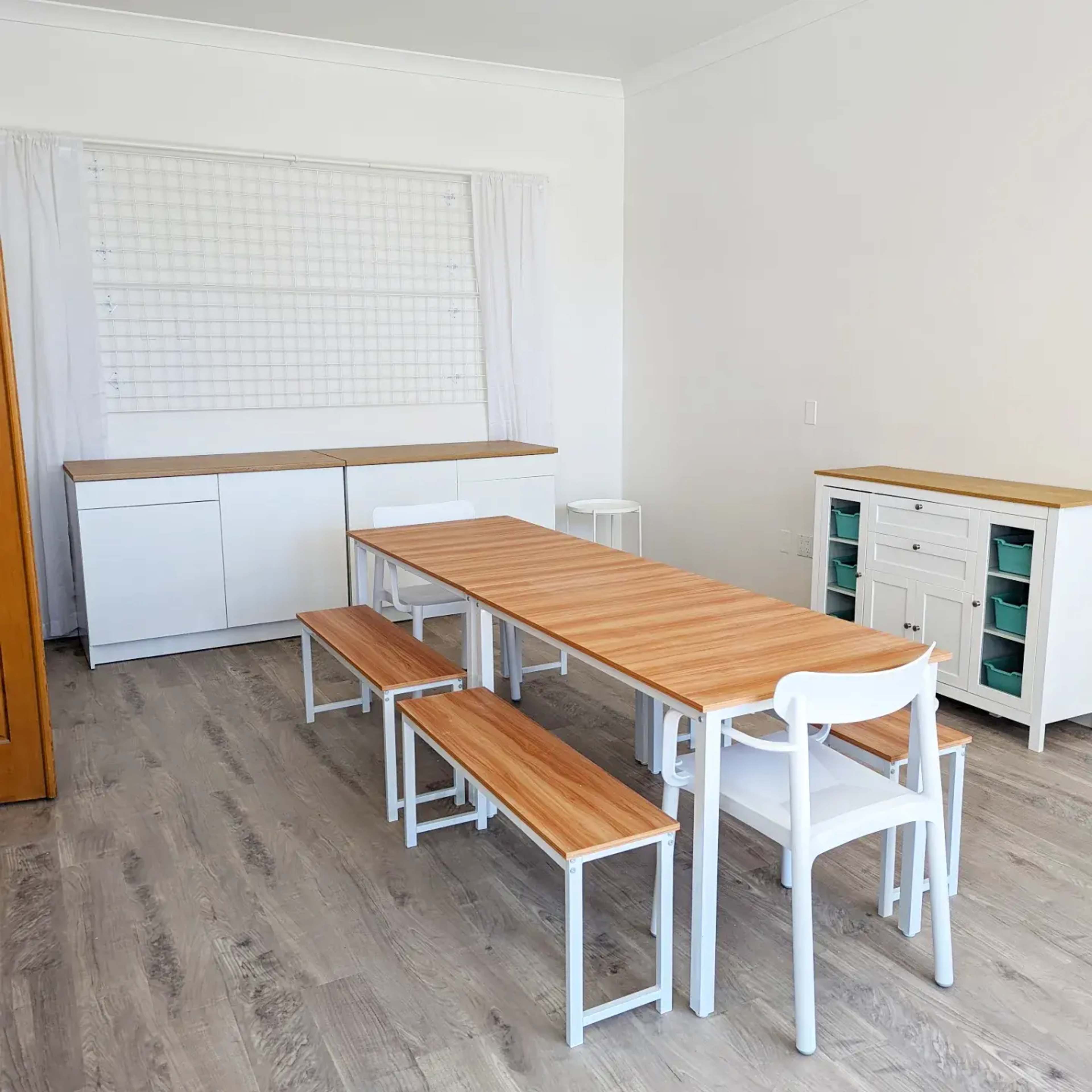A spacious, minimalistic room features a long wooden dining table with white benches and chairs, alongside white cabinets and light-colored walls.