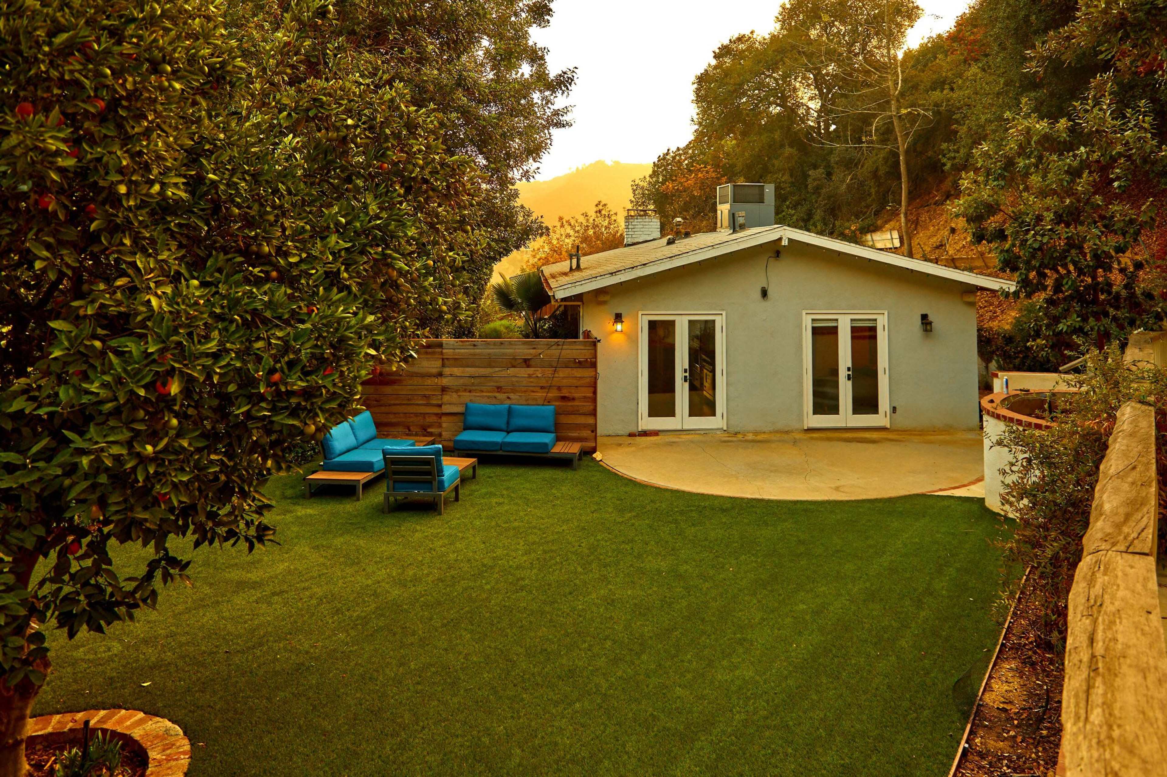 A small house with blue patio furniture sits on a green lawn surrounded by trees.