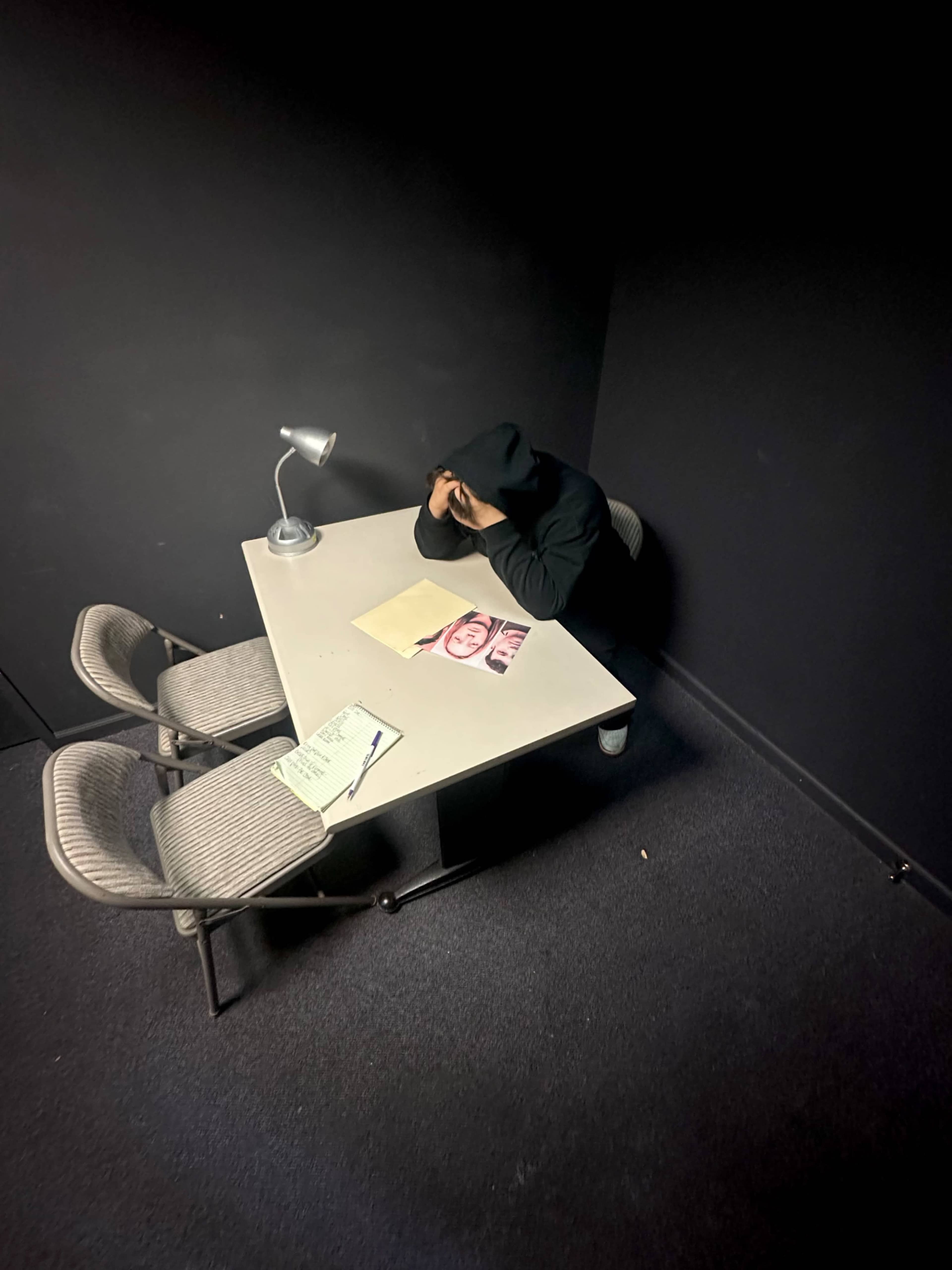 A person wearing a hoodie sits at a table in a dimly lit room, with a lamp and papers on the table.