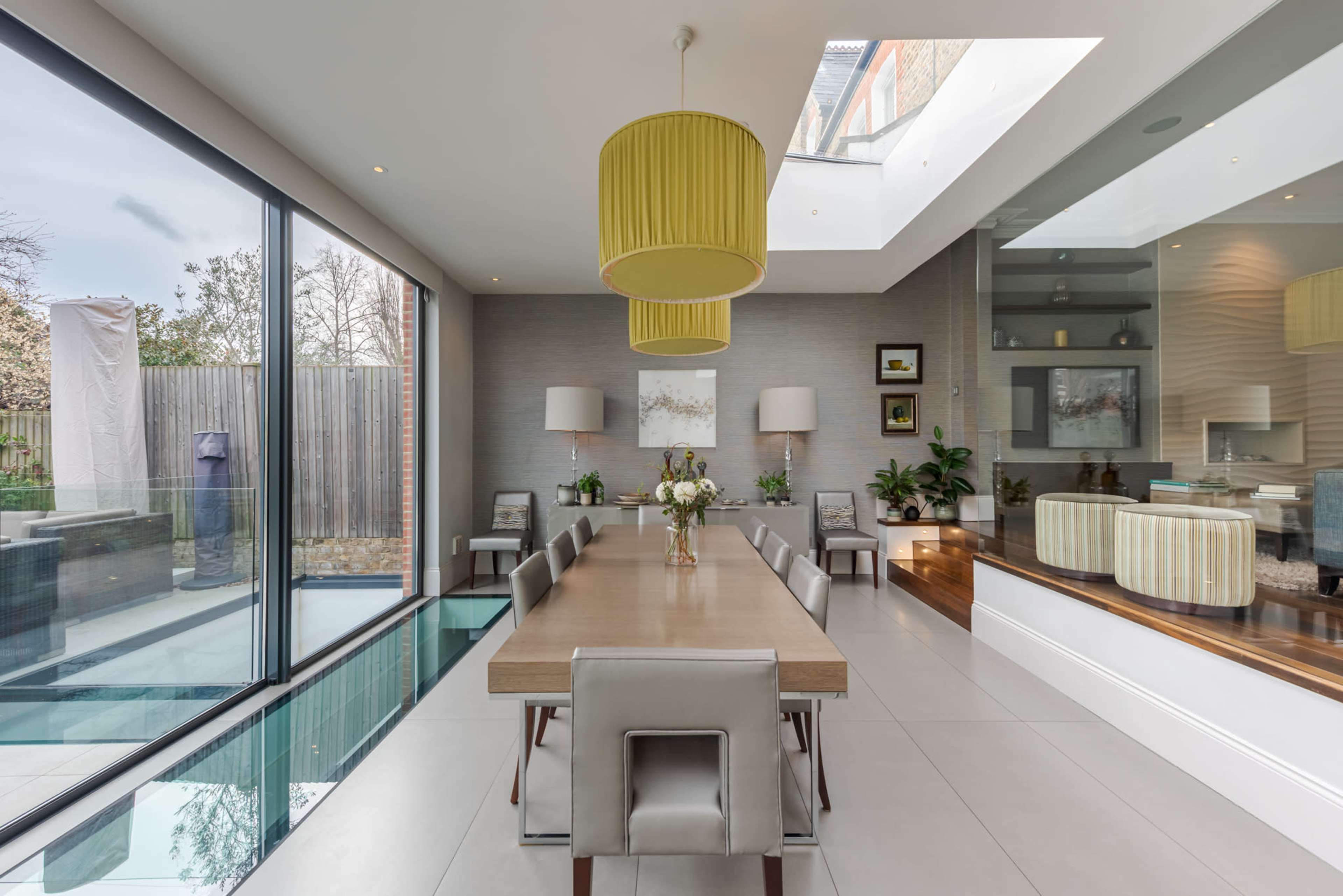 A modern dining room with a long wooden table surrounded by upholstered chairs, large windows allowing natural light, and a glass floor section revealing the area below.