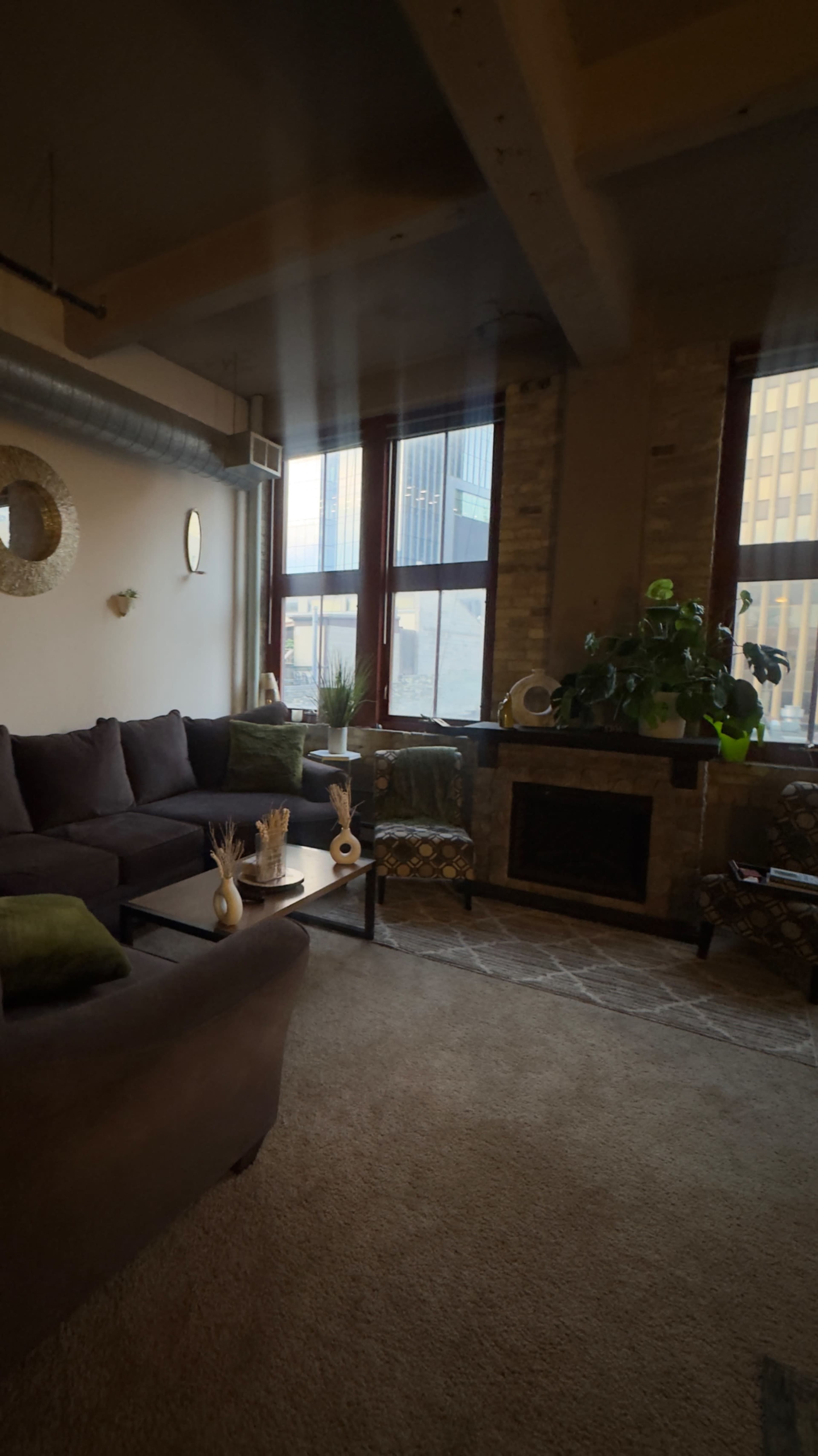 The image shows a cozy living room with large windows, a brown sectional sofa, and a fireplace beside potted plants and decorative items on a coffee table.