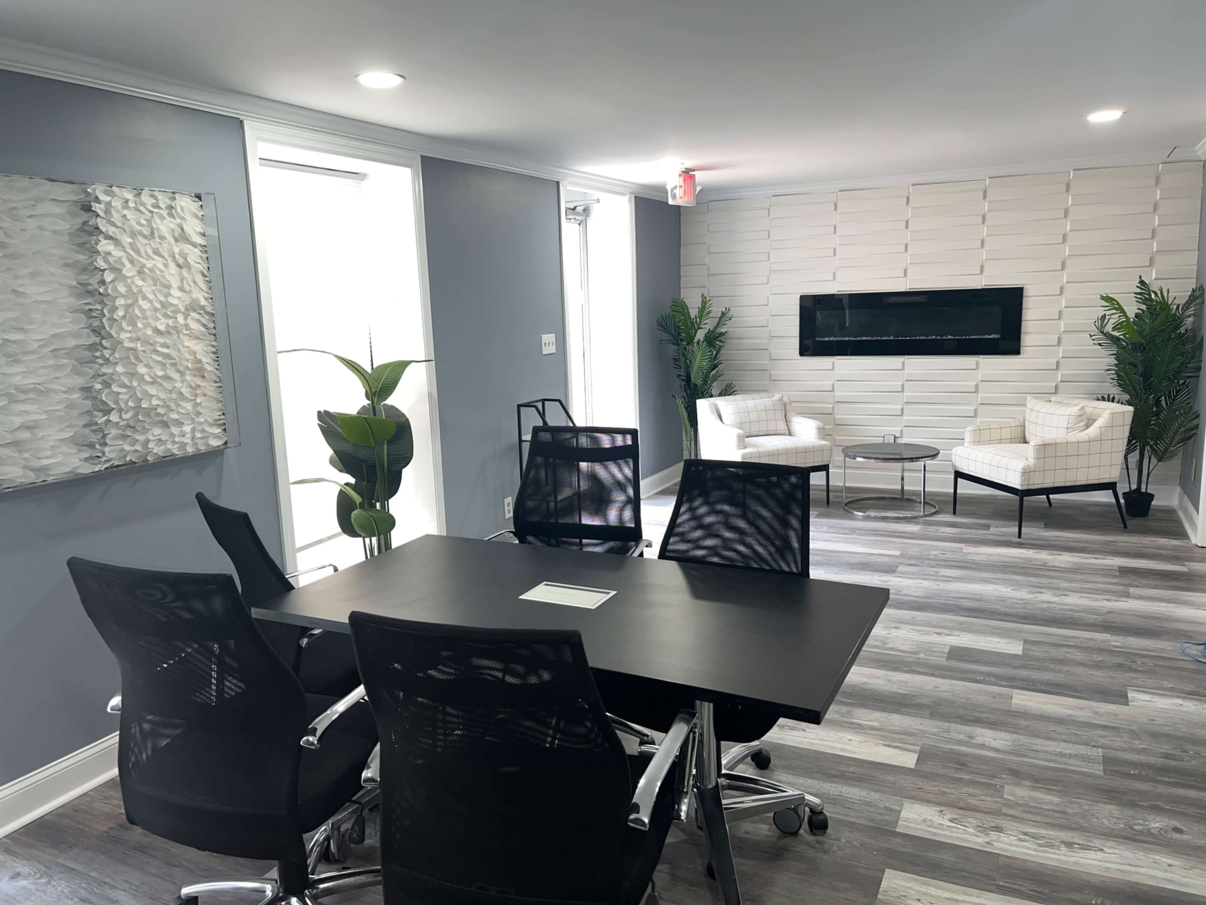 A modern office space with a conference table and four ergonomic chairs, flanked by two plants, and a seating area featuring two armchairs and a wall-mounted fireplace.