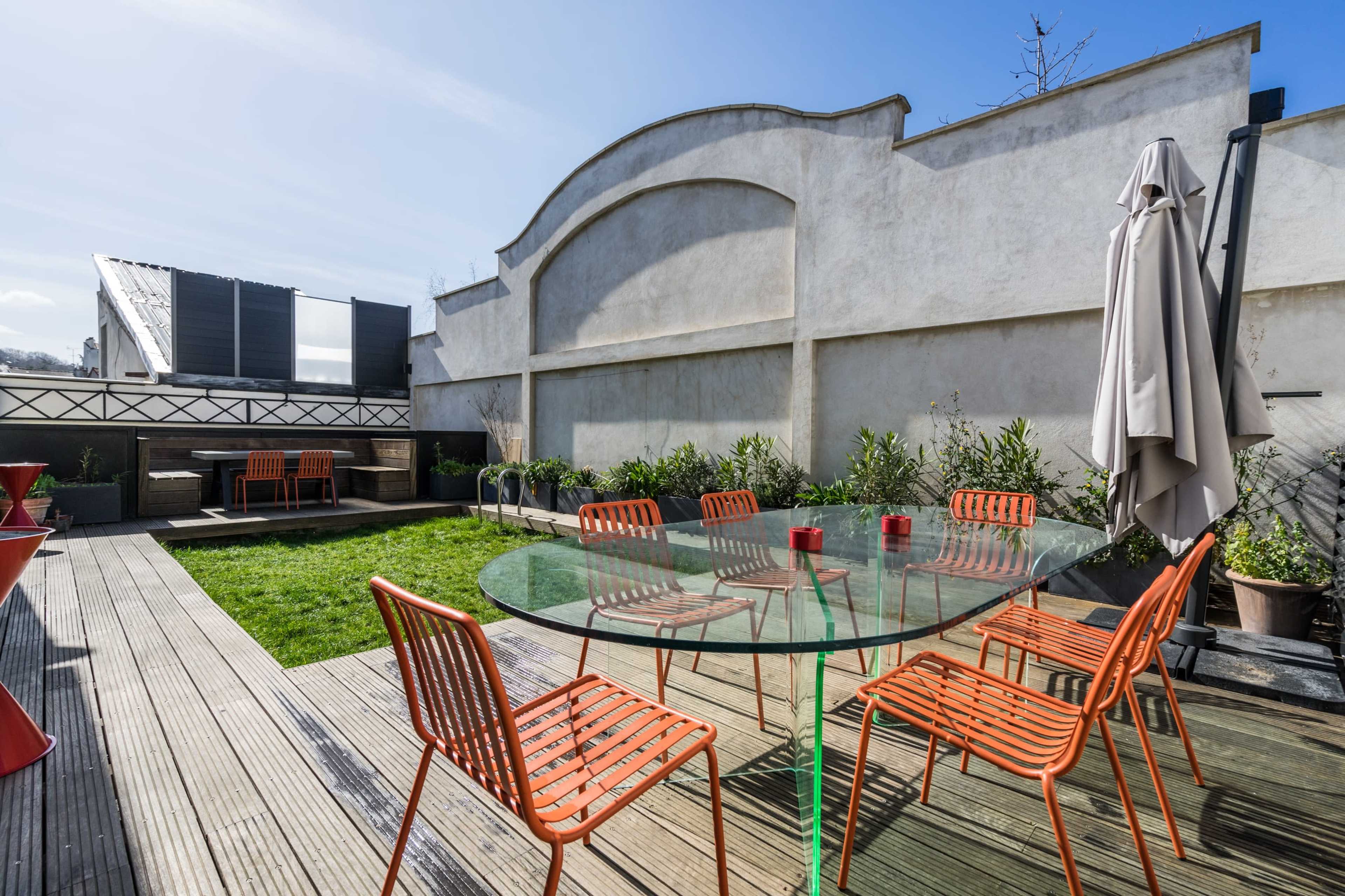 A rooftop terrace features a glass table surrounded by orange chairs, with a green lawn and seating area against a backdrop of a wall.