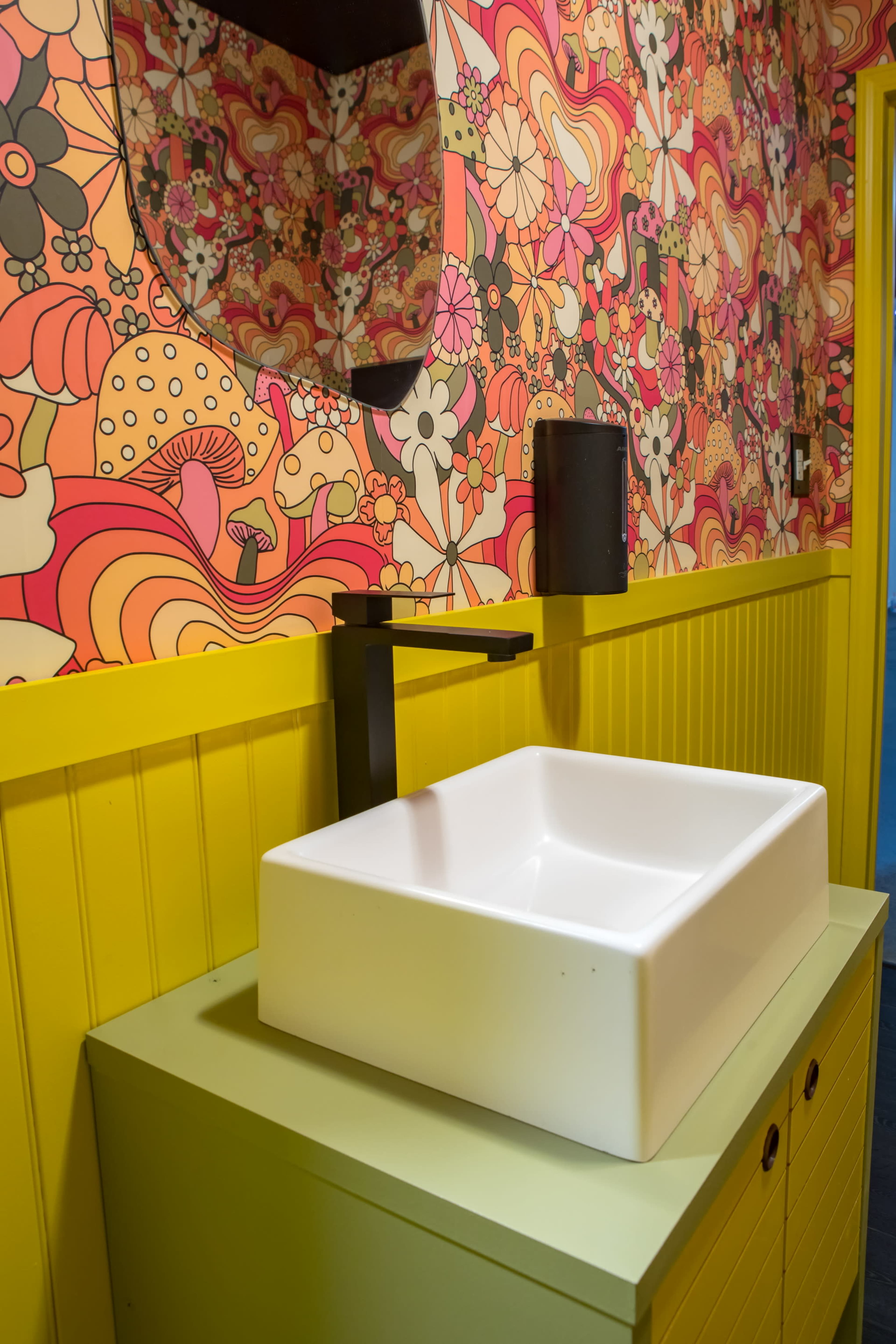 A modern bathroom with a square white sink, a colorful floral wallpaper, and a bright yellow wall paneling.