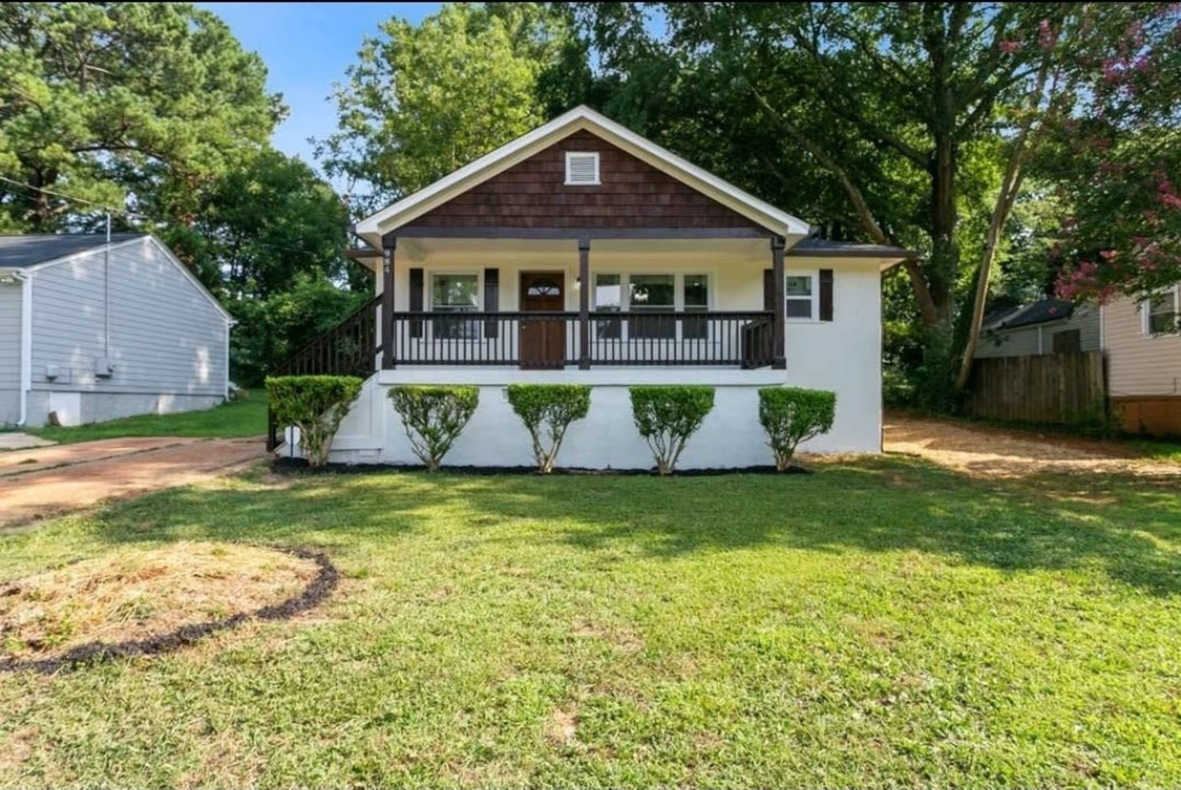 A single-story house with a front porch and a landscaped yard features neatly trimmed shrubs and is surrounded by trees.