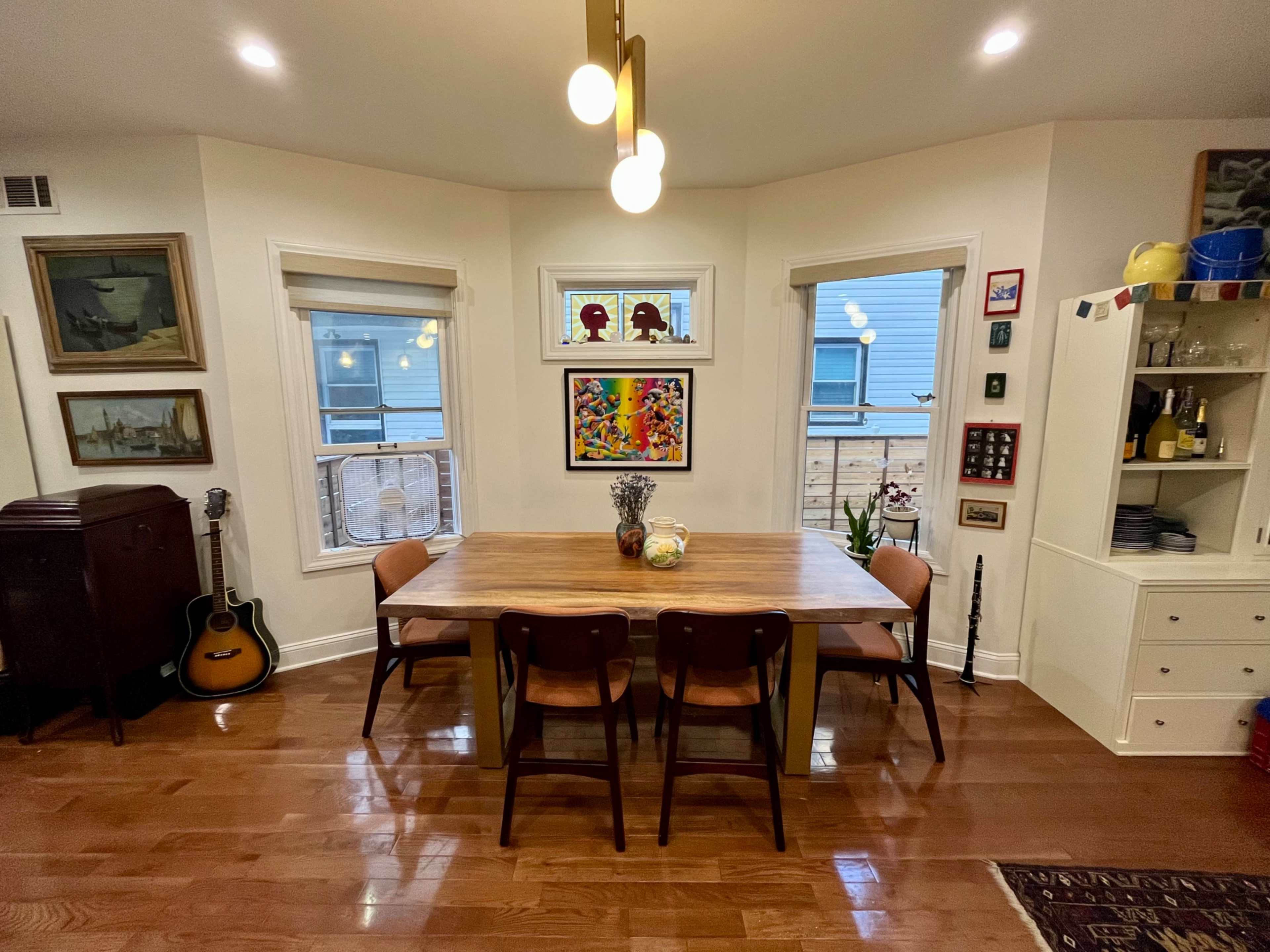 A wooden dining table with four chairs is centered in a well-lit room featuring art on the walls and a guitar resting against a side cabinet.