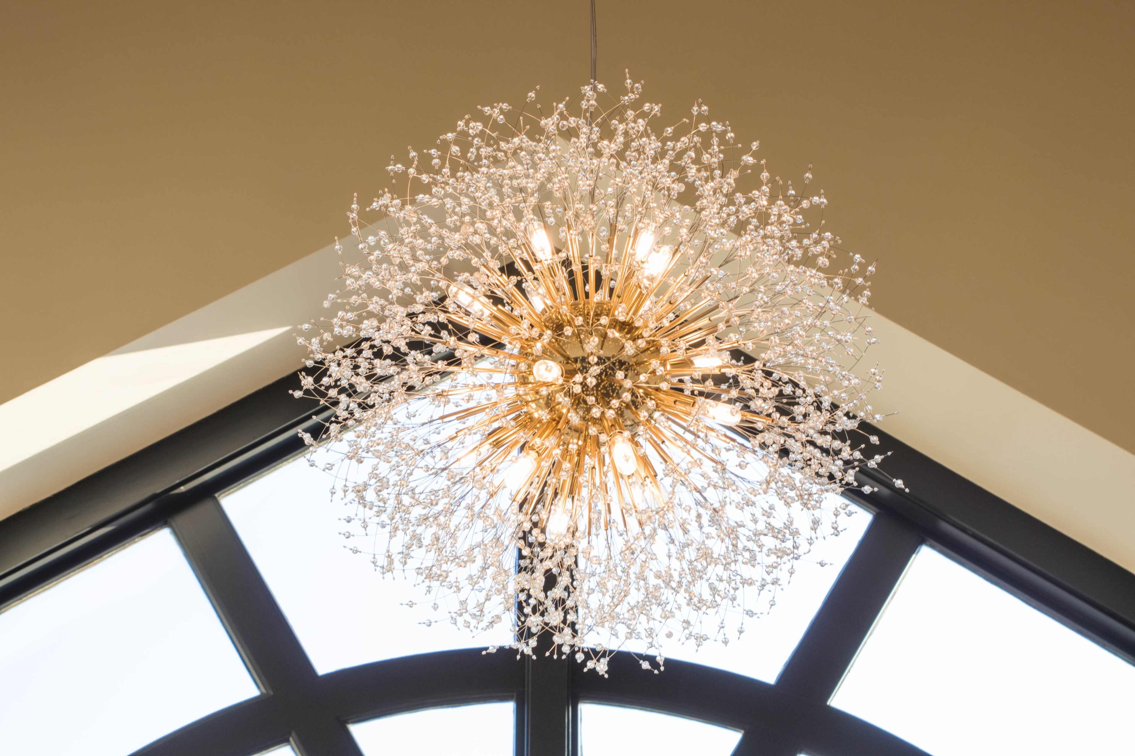A modern chandelier with a spherical design and numerous glass elements hangs from a ceiling in front of large, arched windows.