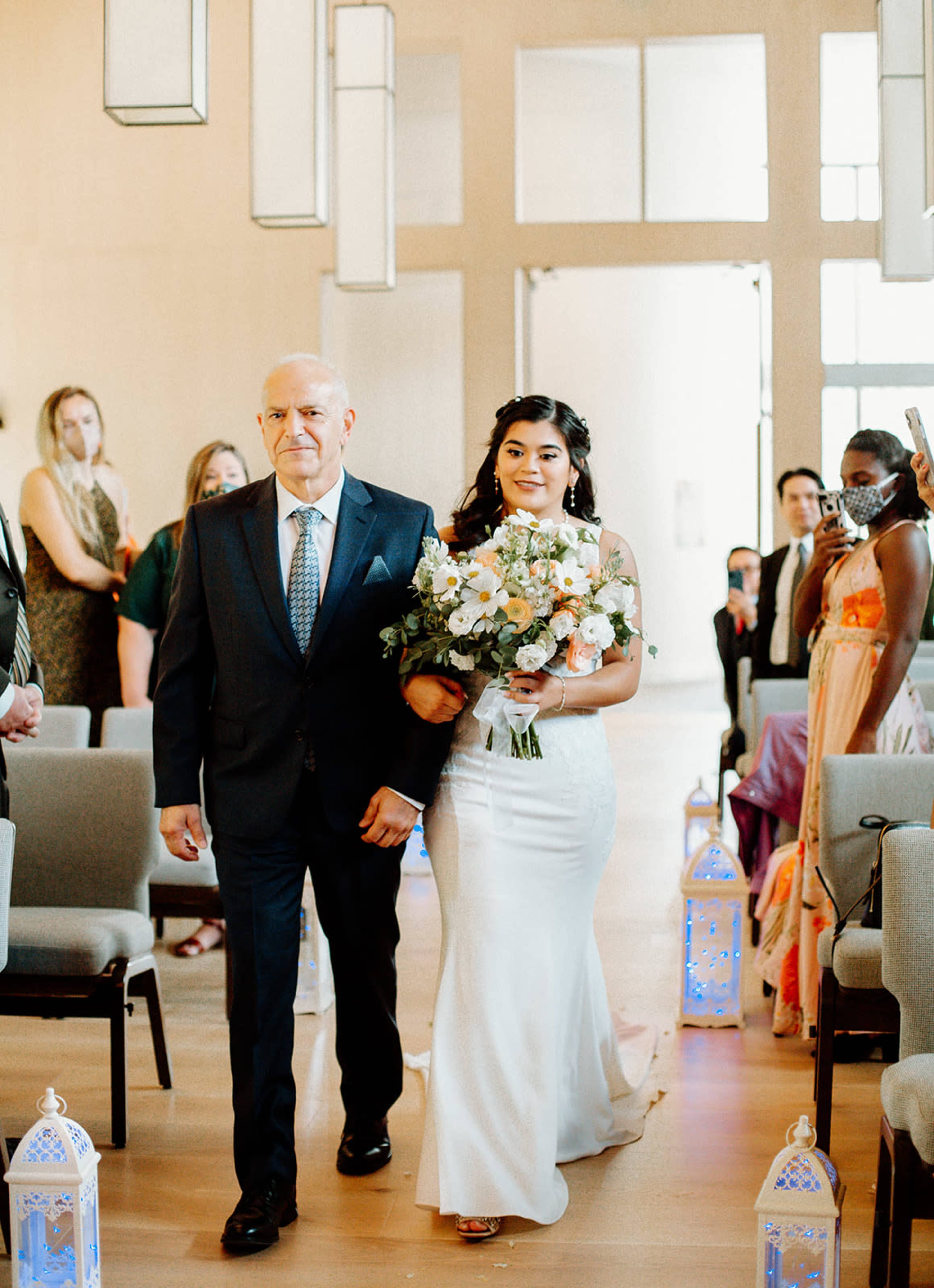 A bride in a white gown walks down an aisle accompanied by her father, as guests look on in a well-lit venue.