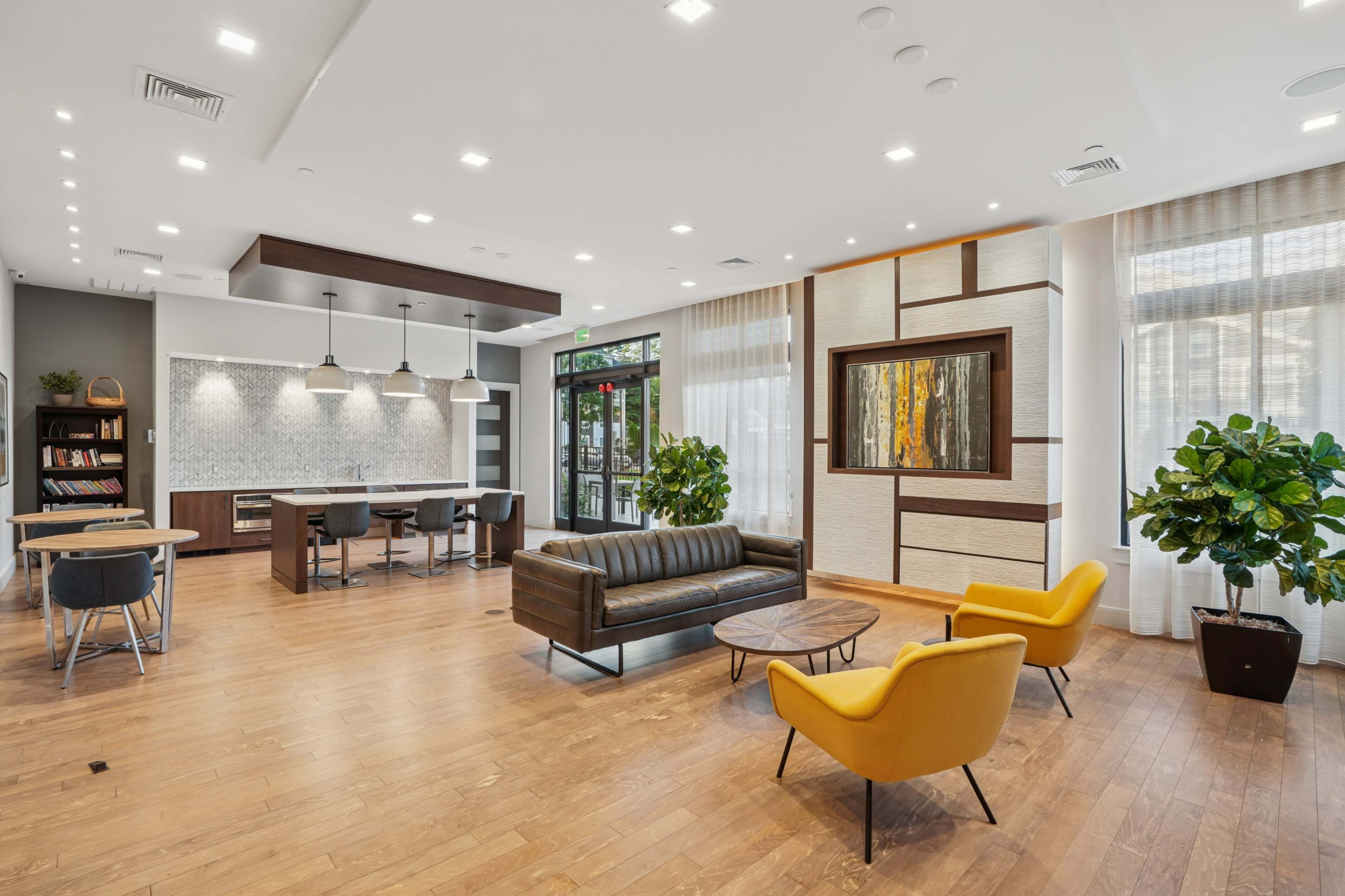 The image shows a modern lounge area featuring a leather sofa, two yellow chairs, a wooden coffee table, and a kitchenette with bar stools.