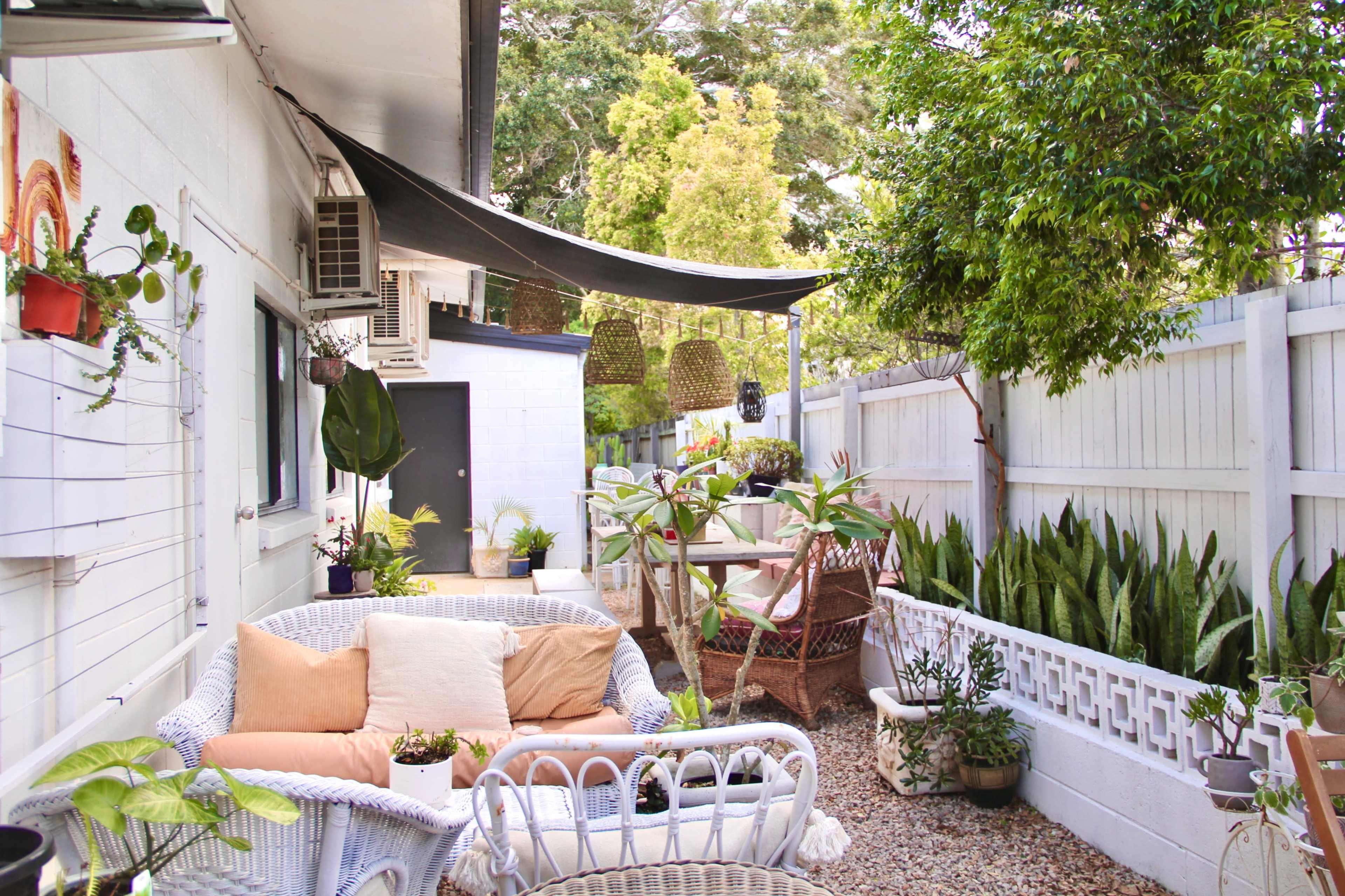 A patio area with wicker furniture, potted plants, and a shaded dining table surrounded by greenery and a wooden fence.