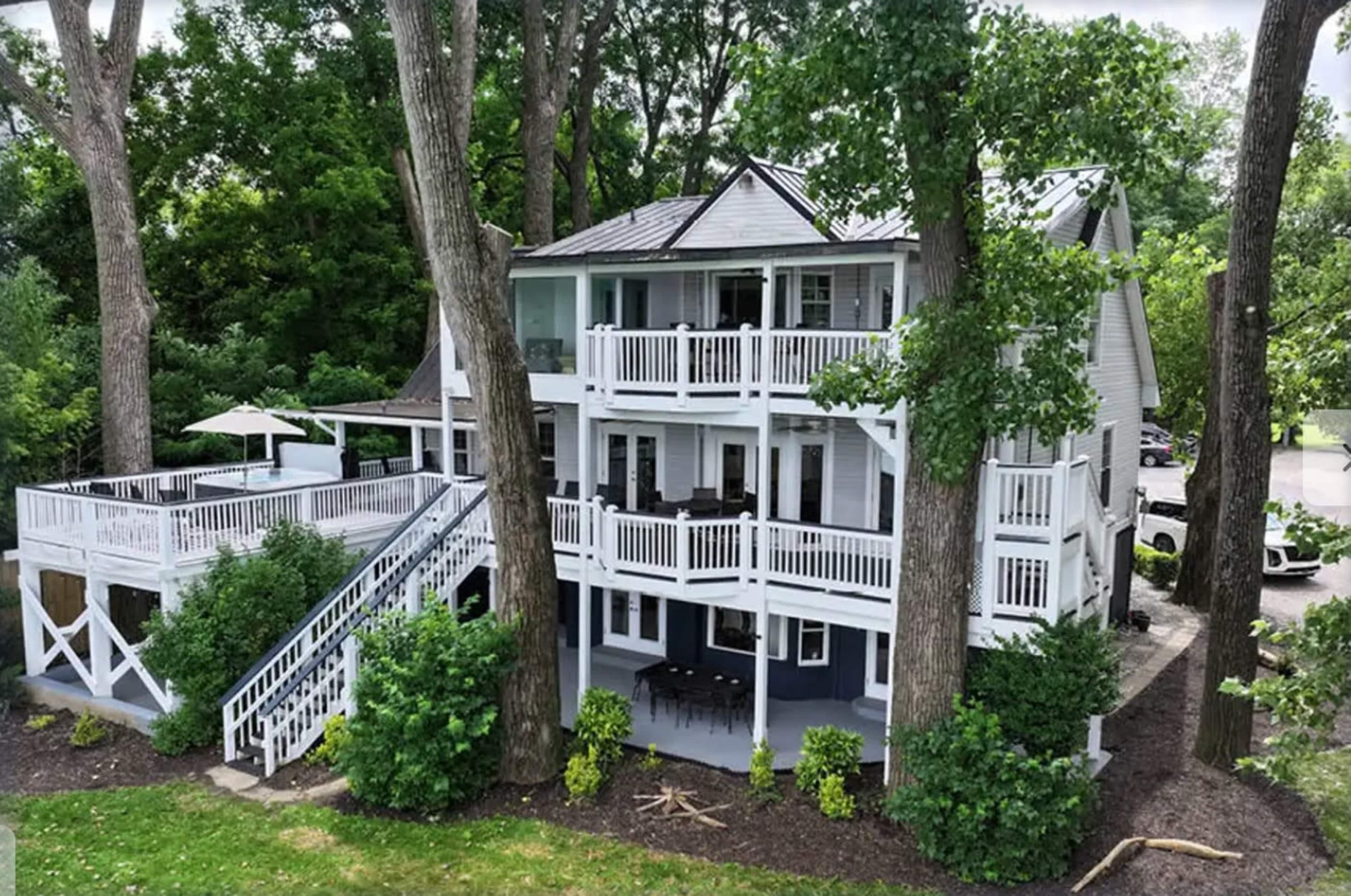 A three-story white house with multiple balconies is surrounded by large trees and greenery.
