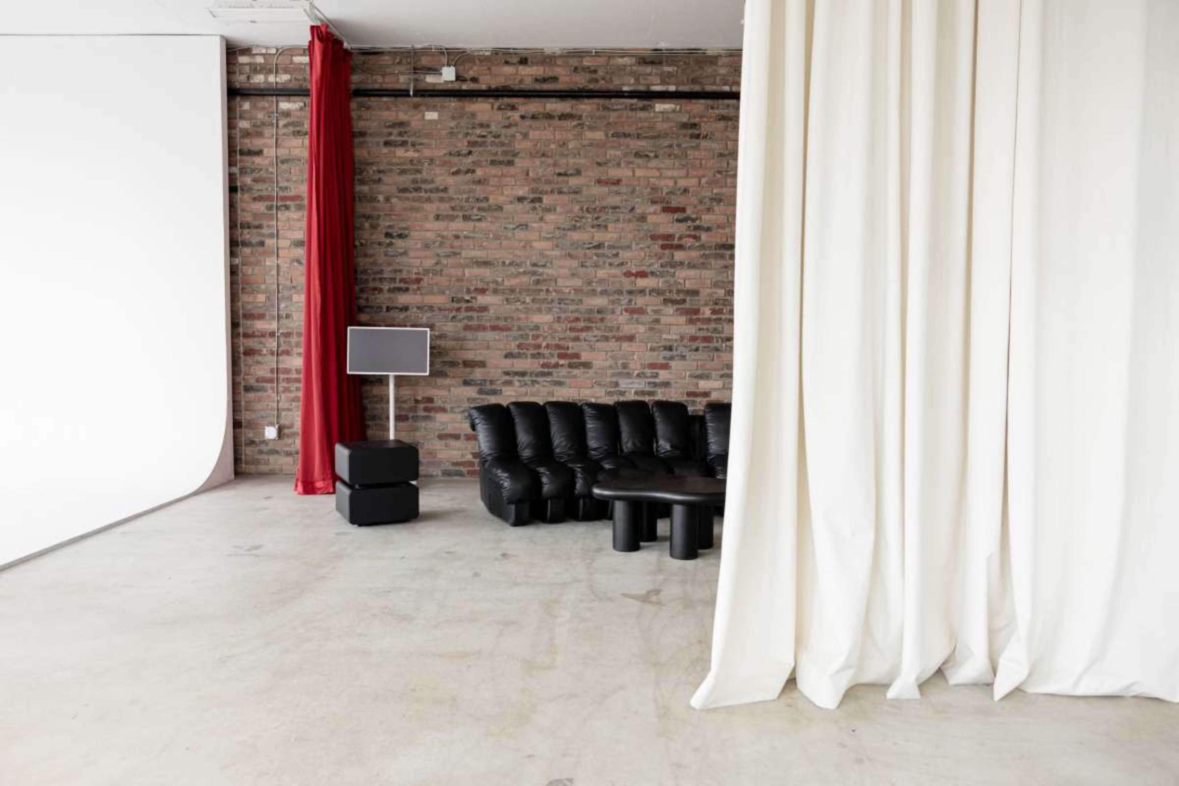 The image shows a studio space with exposed brick walls, a black leather couch, a black coffee table, and a white backdrop draped alongside a red curtain.