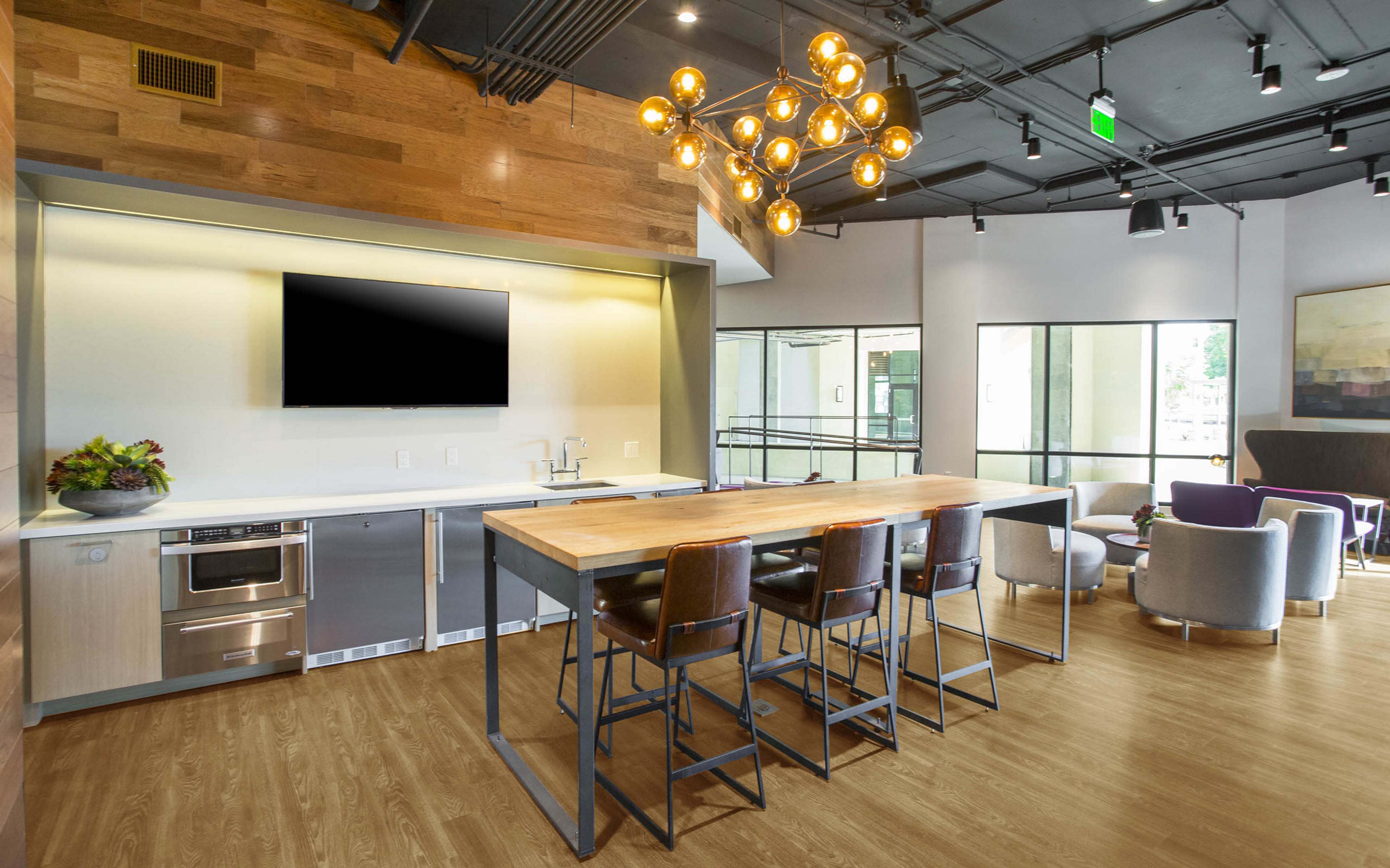A modern kitchenette area featuring a large wooden table with high chairs, an LED television mounted on the wall, and large windows allowing natural light to enter.