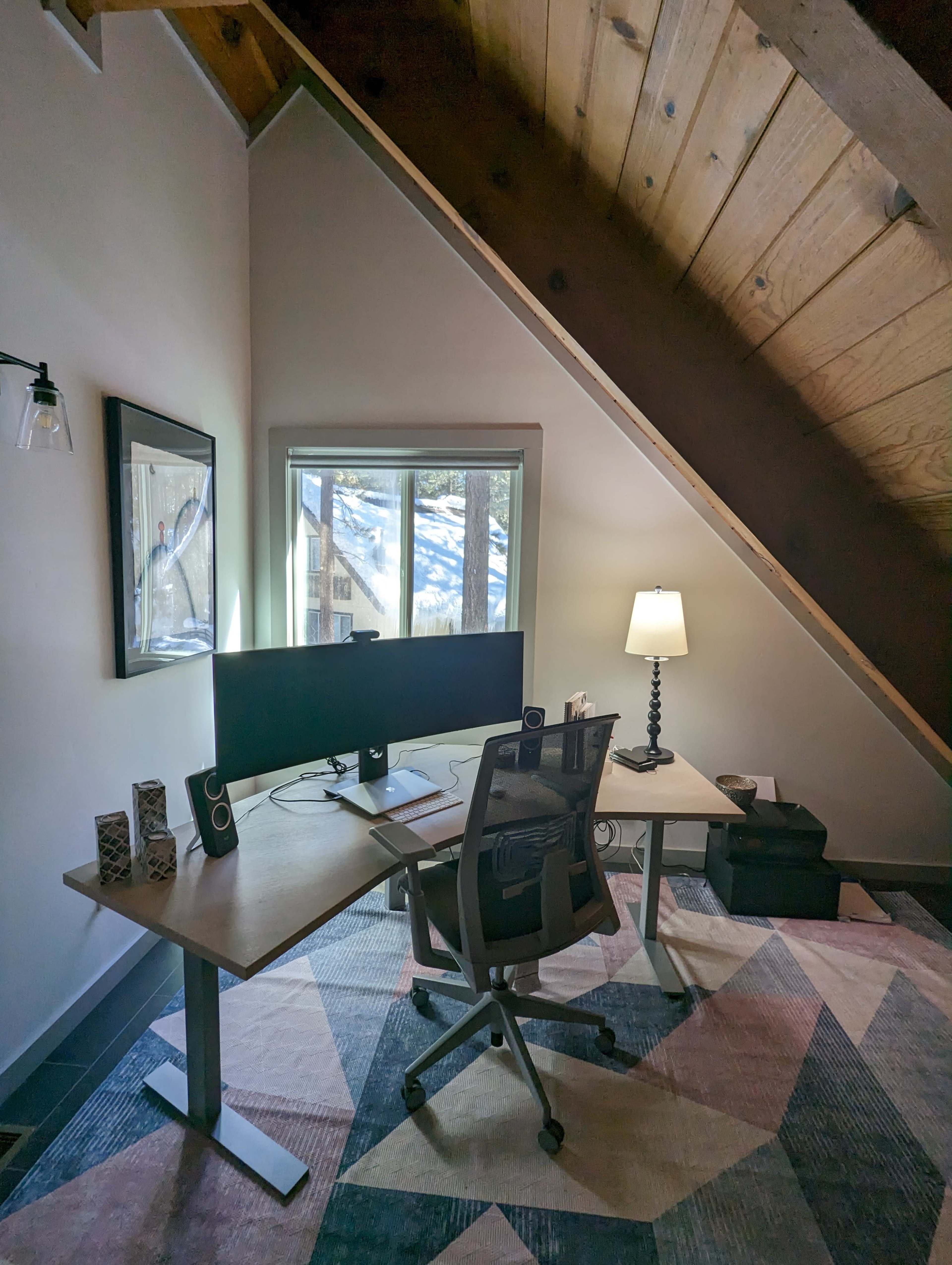 A modern home office situated under a sloped wooden ceiling, featuring a desk with a computer monitor, a comfortable chair, and a lamp next to a window displaying snow-covered scenery outside.