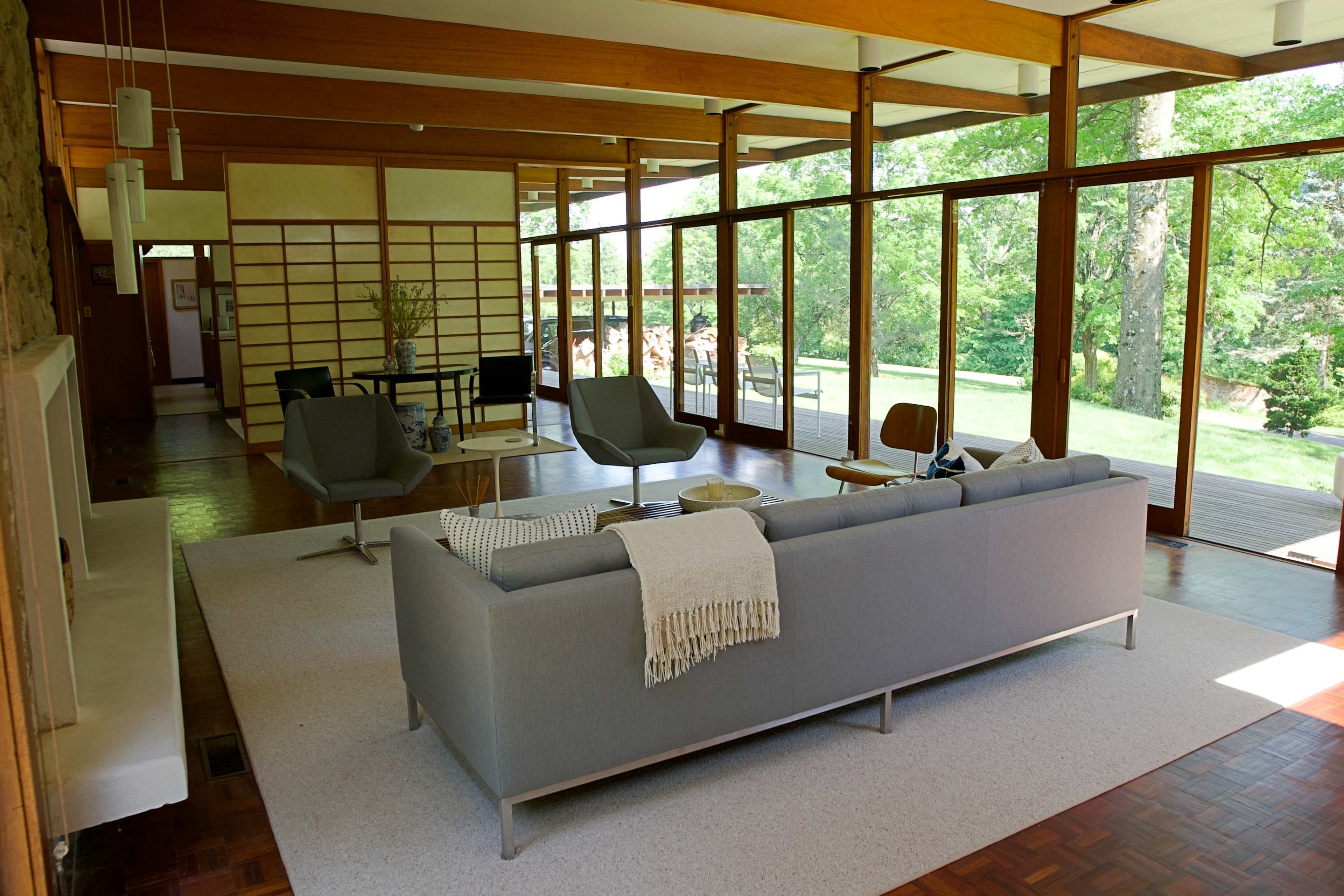 A modern living room features a gray sofa, wooden furniture, and large windows that overlook a green landscape.