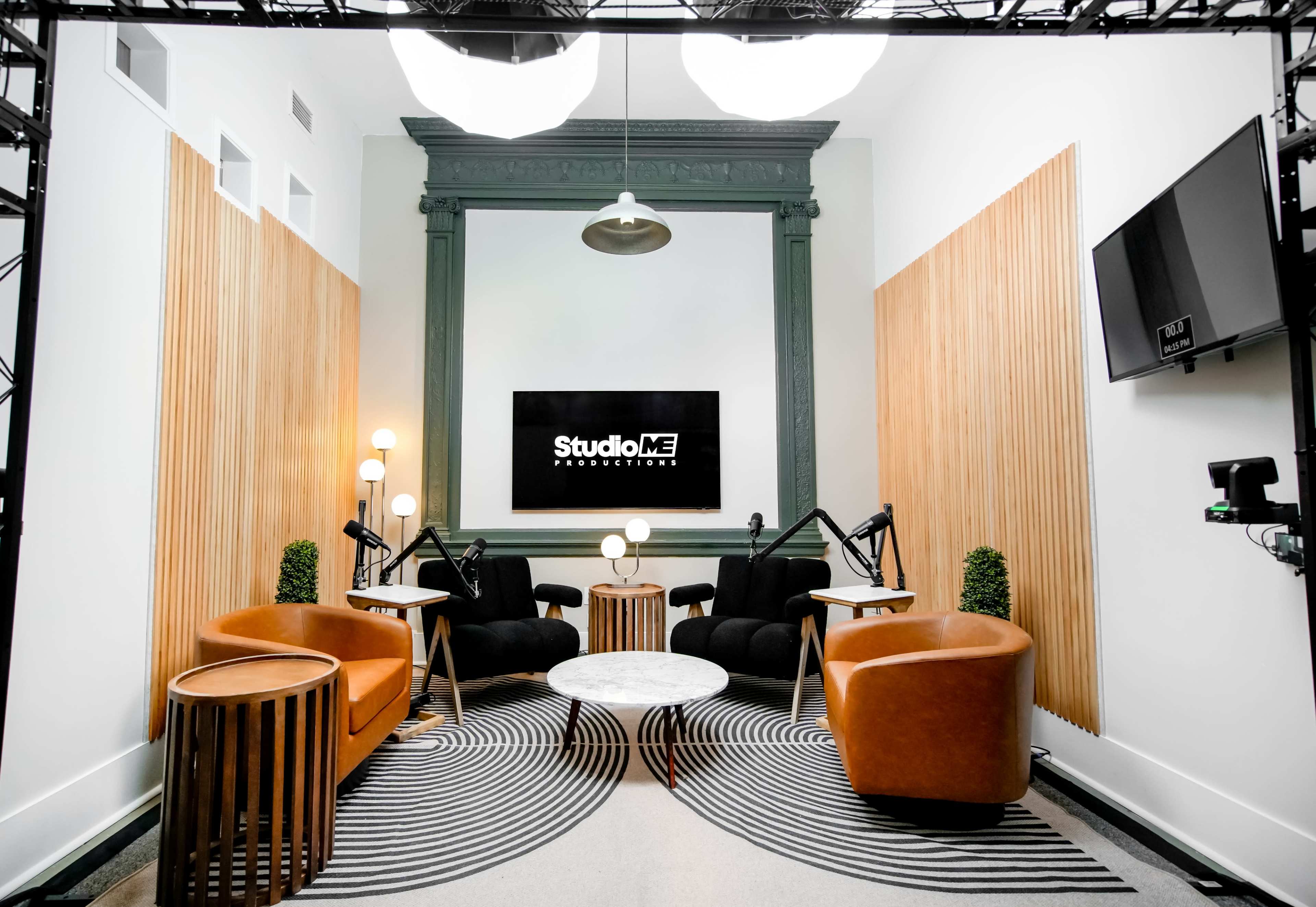 The image shows a modern studio space with two brown sofas, a round coffee table, and a large screen displaying "StudioME Productions" on the wall.