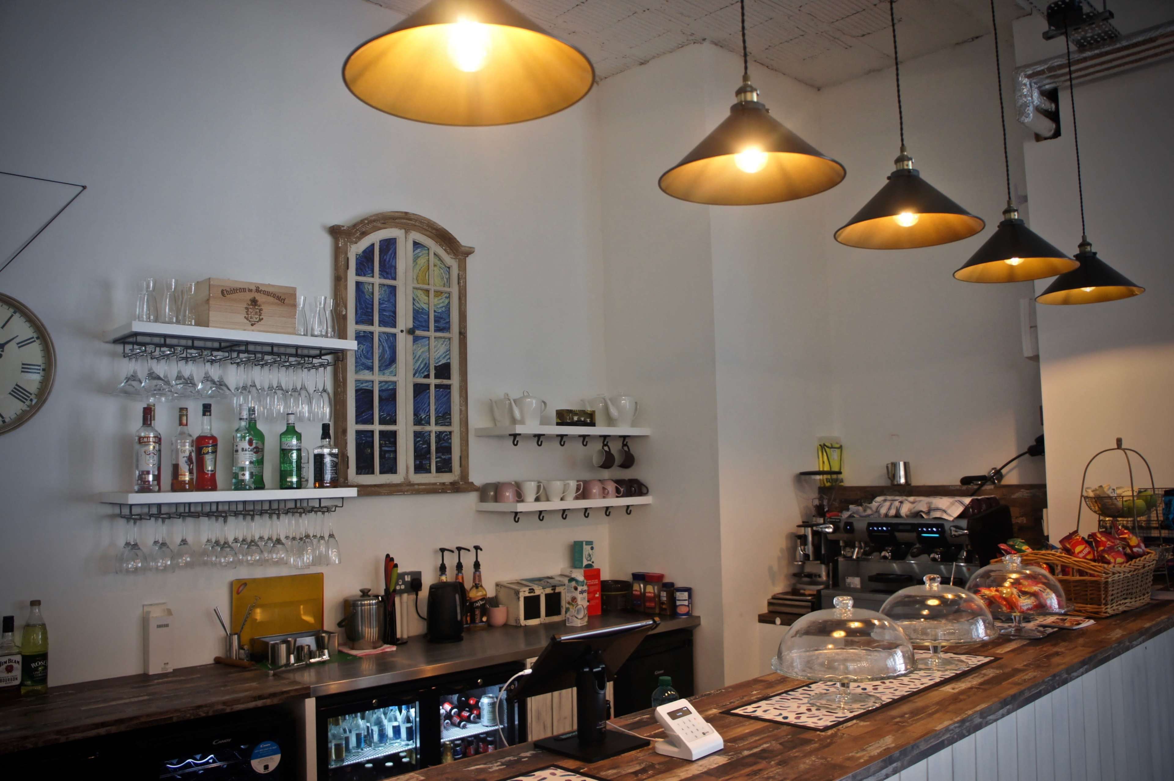 A café interior with a wooden counter, multiple shelves holding glassware and bottles, and pendant lights hanging overhead.