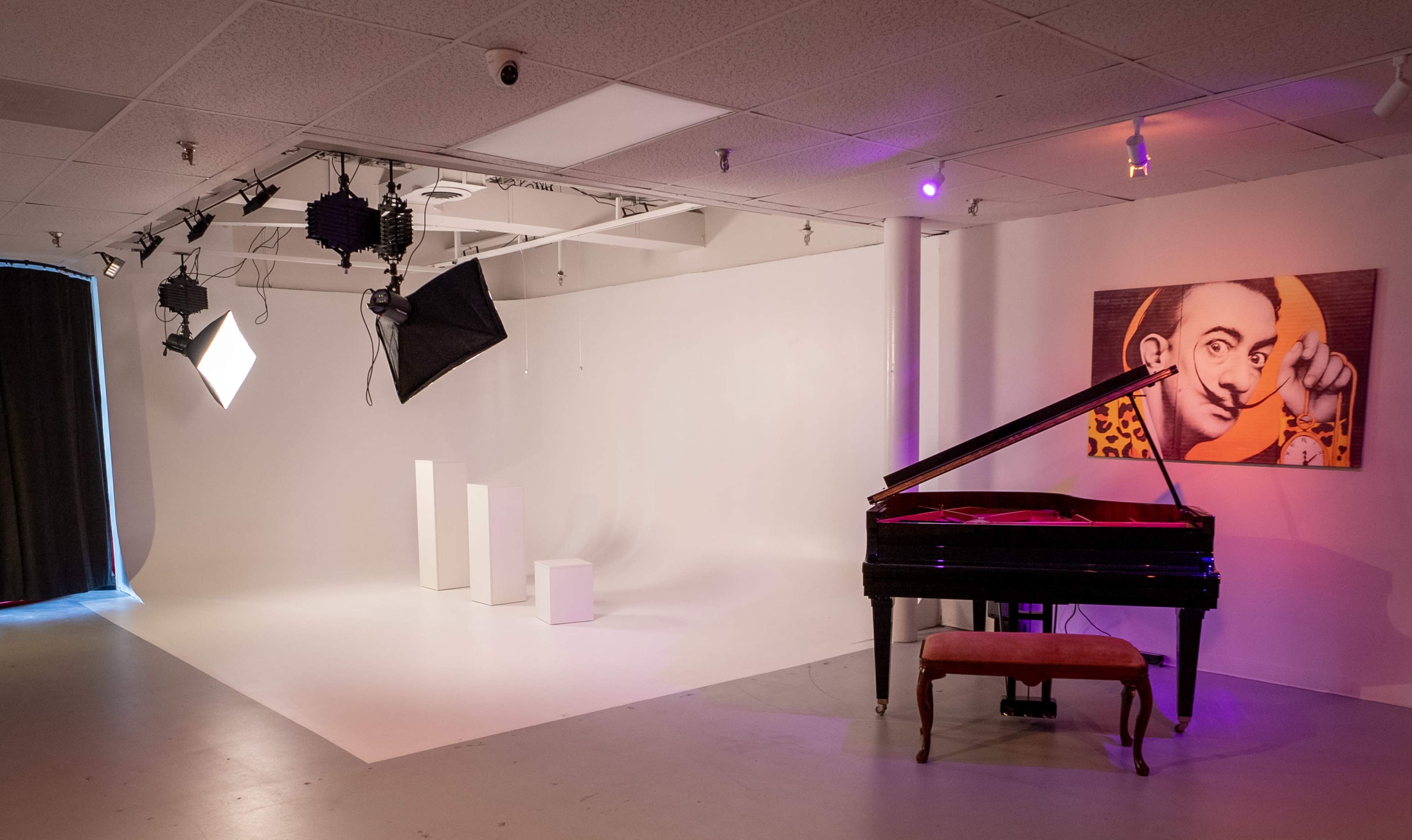 The image shows a spacious studio with a grand piano, a red upholstered bench, three white pedestals, and studio lighting, accompanied by a colorful portrait on the wall.