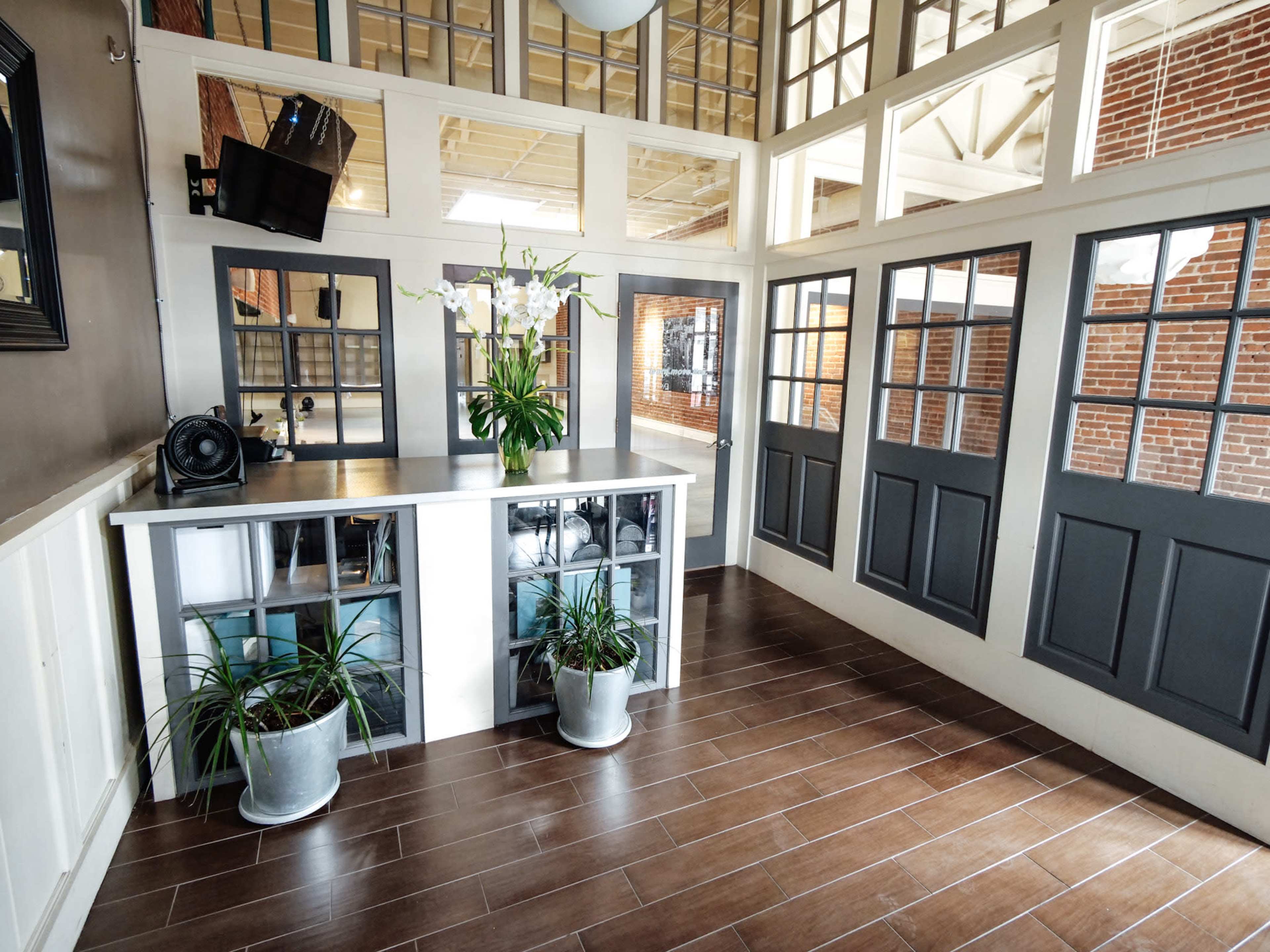 The image shows a spacious reception area with a dark wood floor, a white counter displaying decorative plants, and large windows that allow natural light to fill the room.