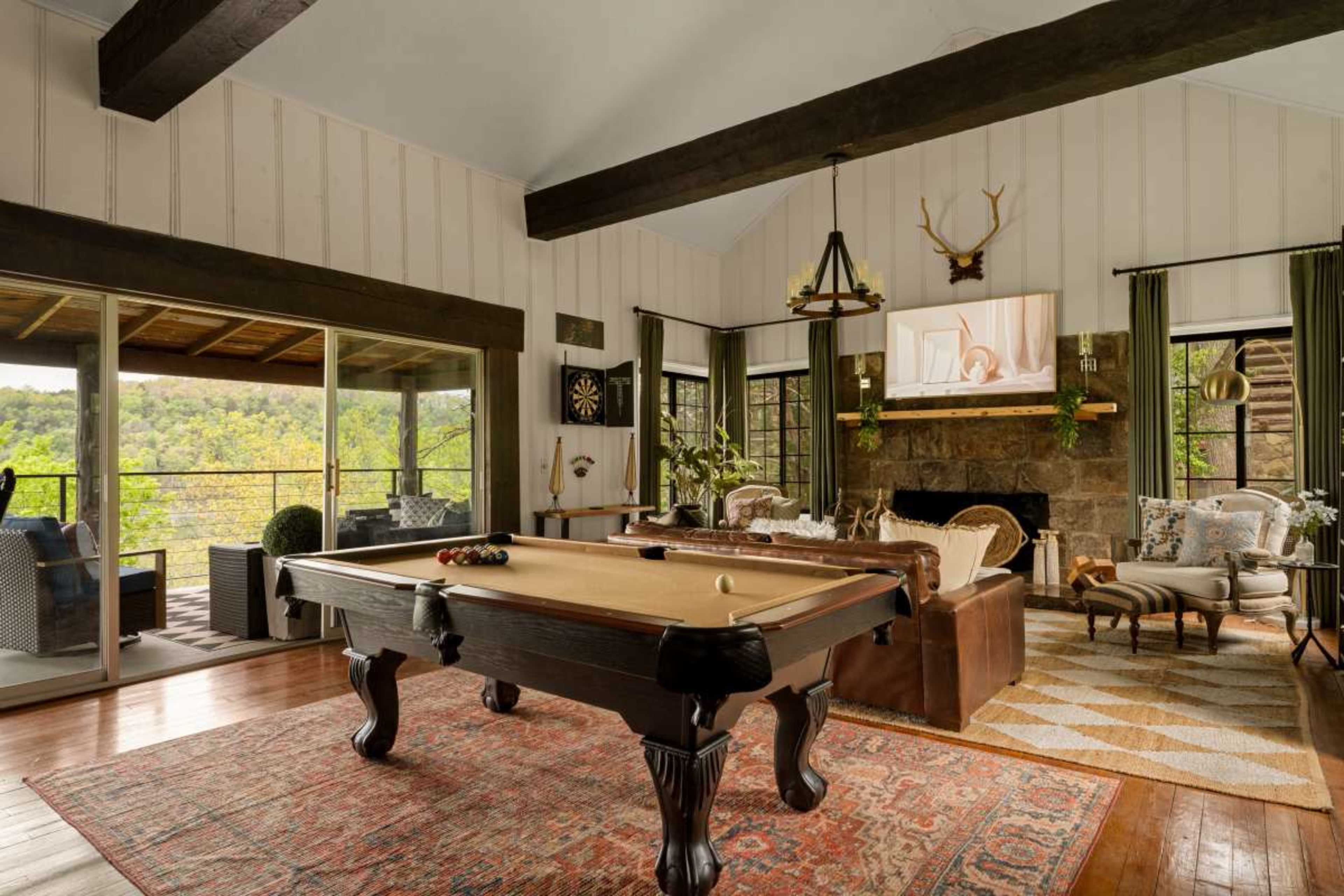 The image shows a cozy living room with a pool table, large windows overlooking a green landscape, and decorative furnishings, including a stone fireplace and a television mounted on the wall.