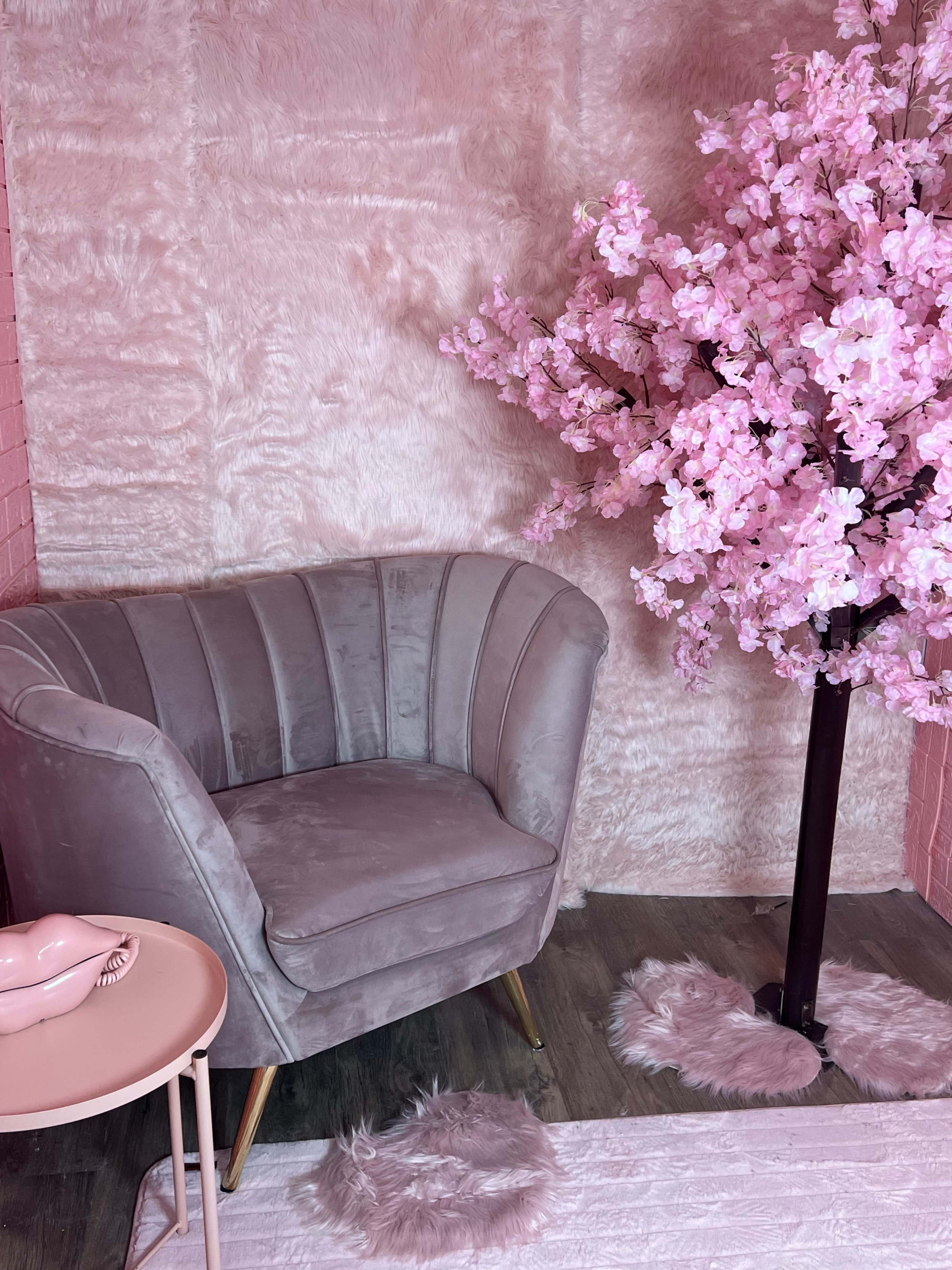 A gray armchair next to a pink side table and a decorative pink cherry blossom tree against a textured pink wall.
