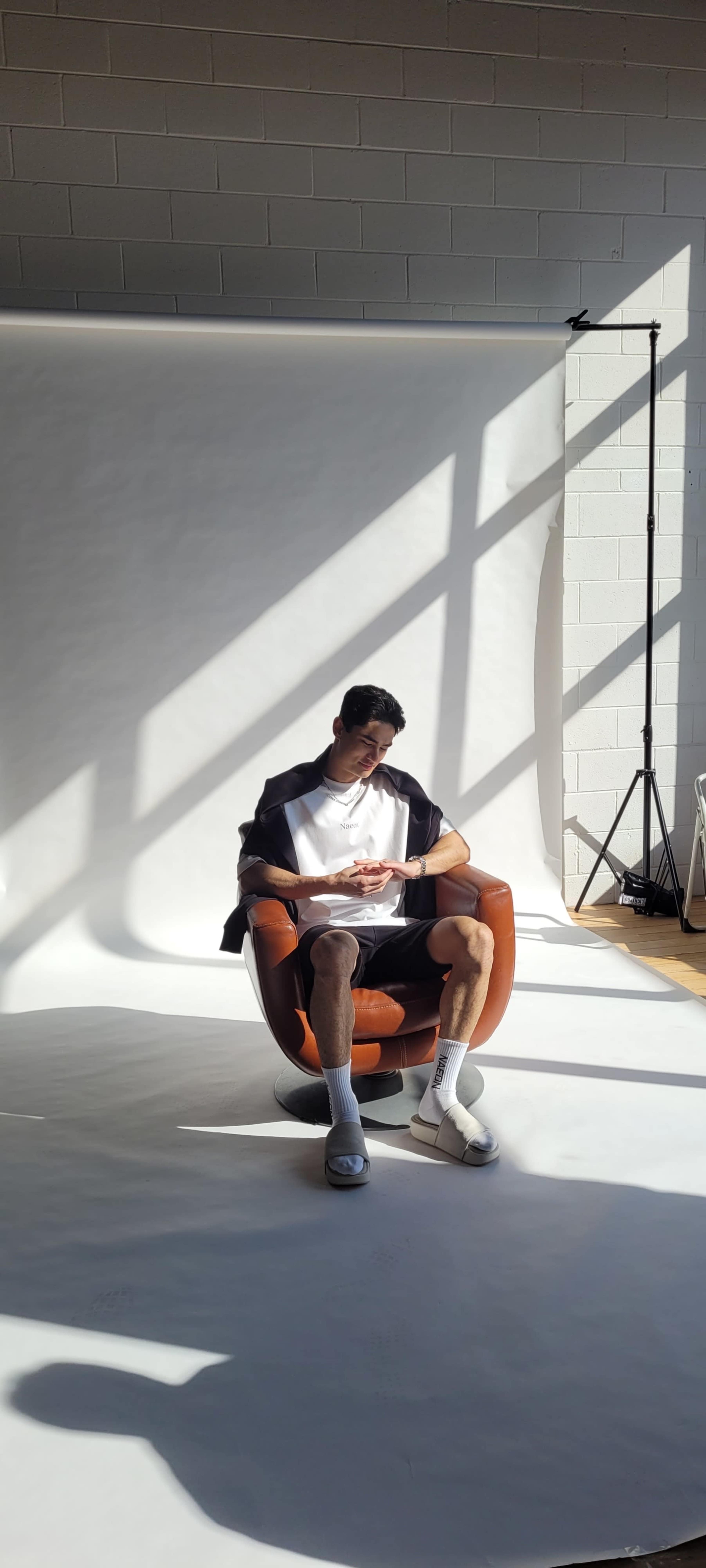 A person sits in a brown chair, reading a book in a well-lit studio with shadows cast by overhead beams.