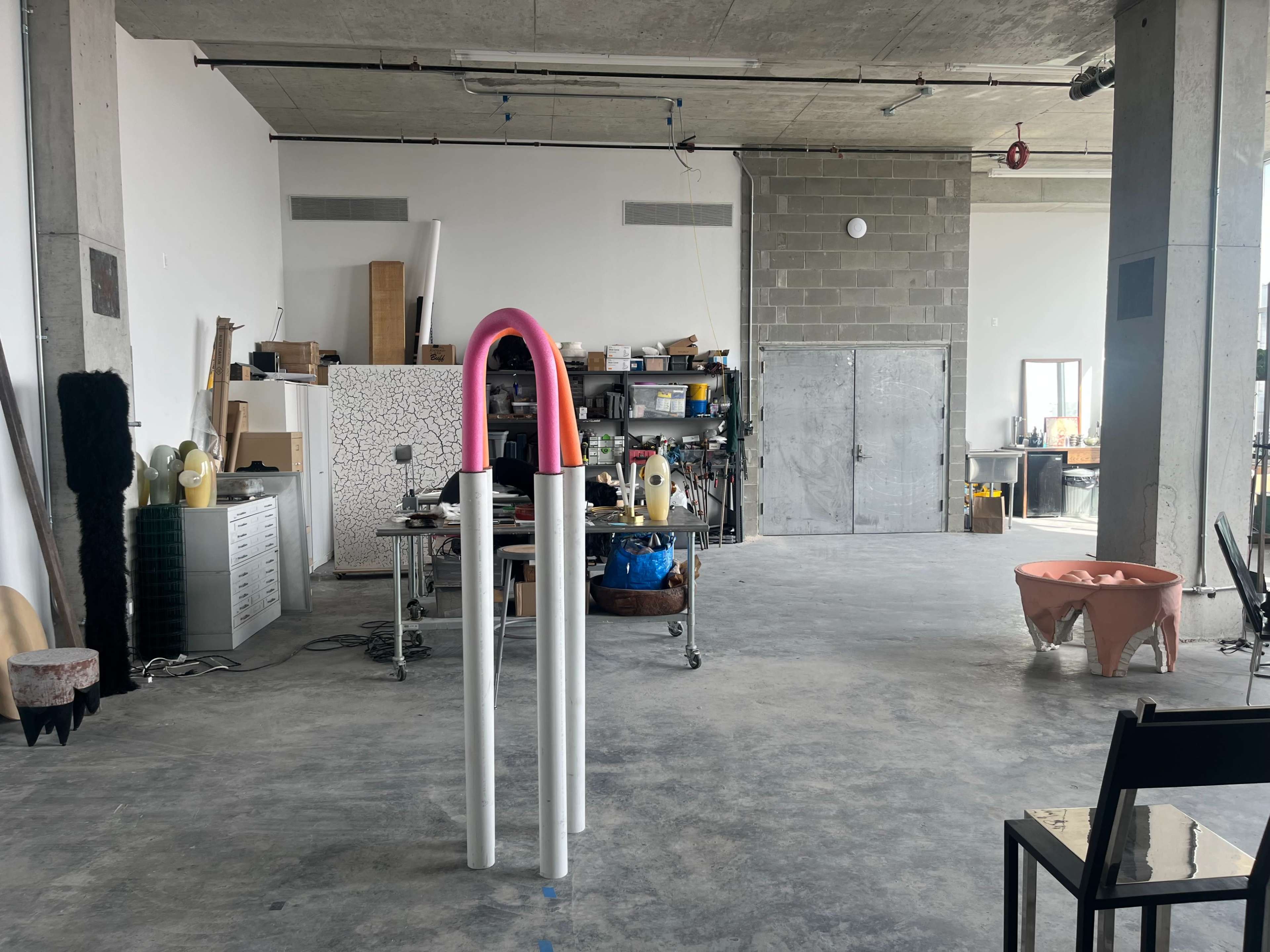 An industrial-style art studio with concrete walls, various tools and materials scattered, and a pink and white sculpture in the foreground.
