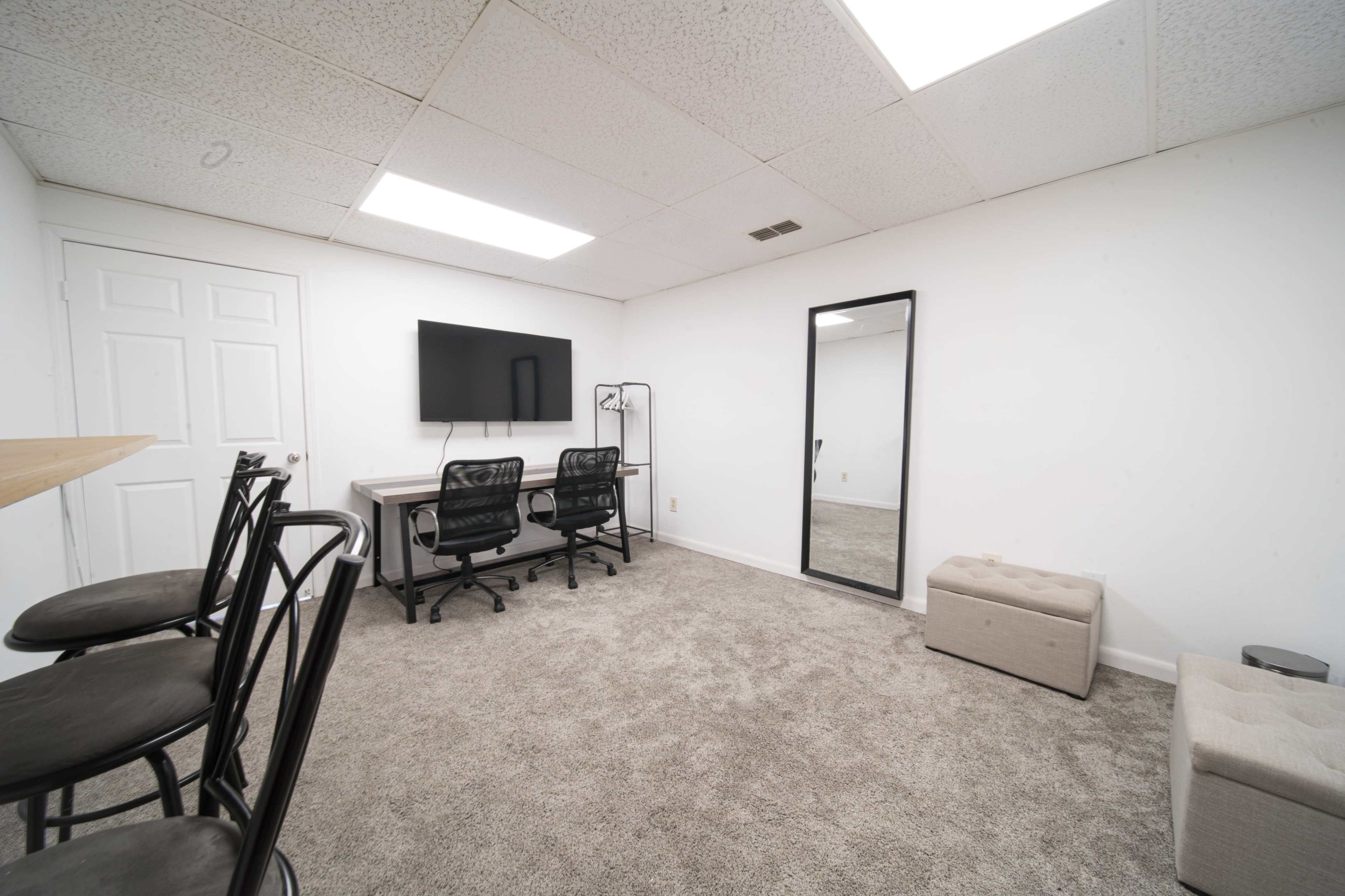 The image shows a simple room with a carpeted floor, featuring a desk, two black office chairs, a large wall-mounted television, a full-length mirror, and a small storage ottoman.