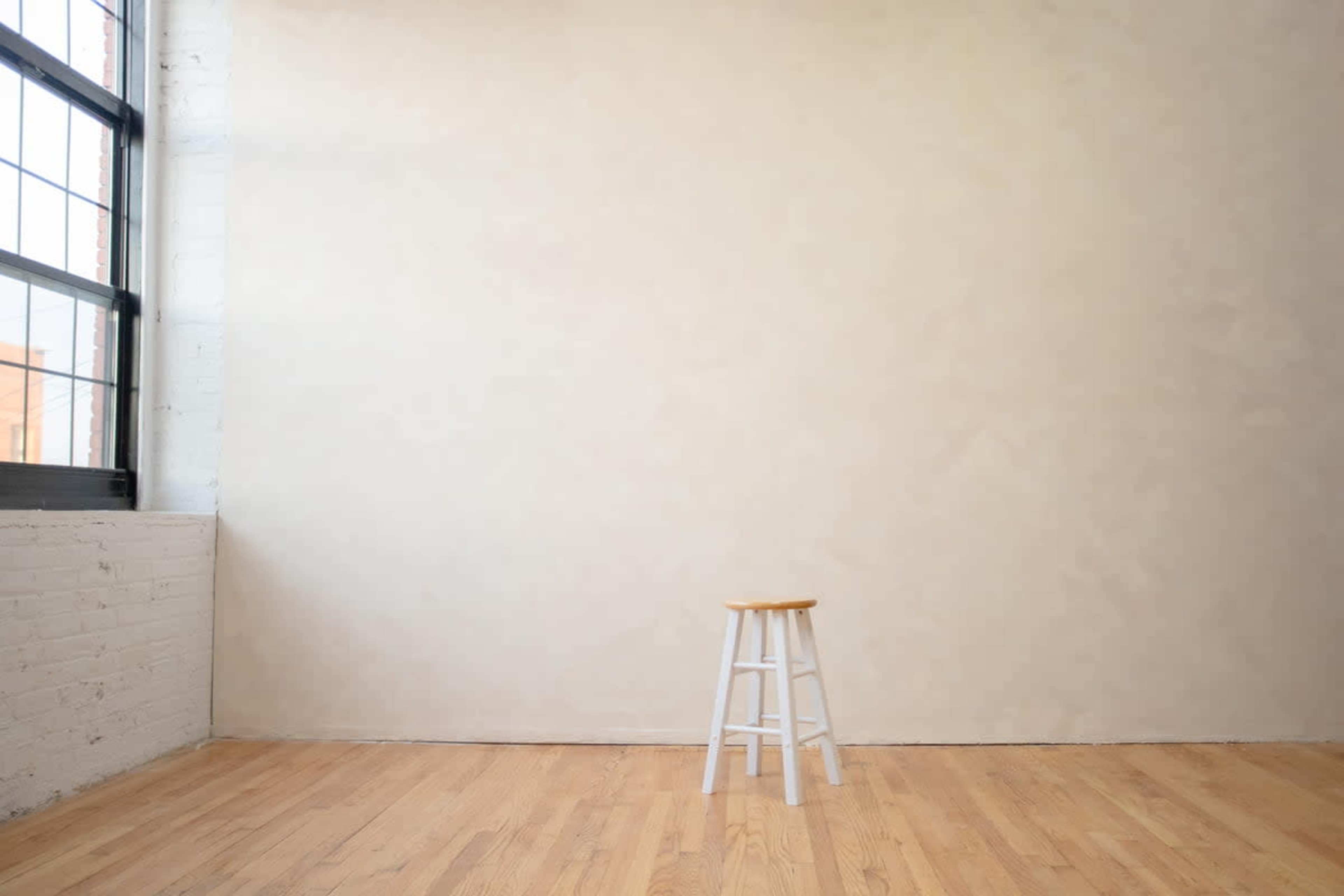 A wooden stool stands alone in an empty room with light-colored walls and a large window.