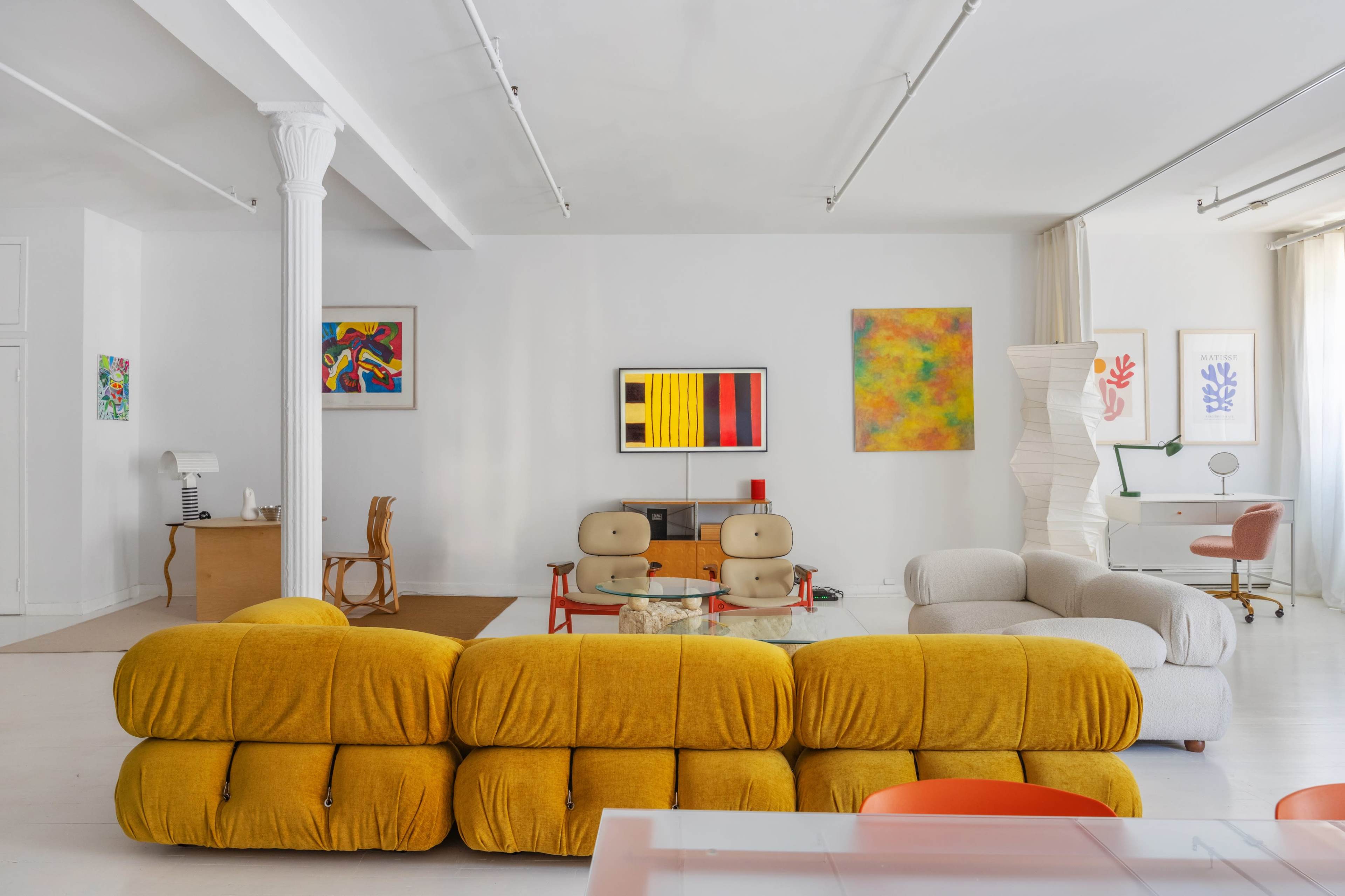 The image shows a modern, minimalistic interior with a yellow sofa, a glass coffee table, two armchairs, and colorful artwork on the walls.