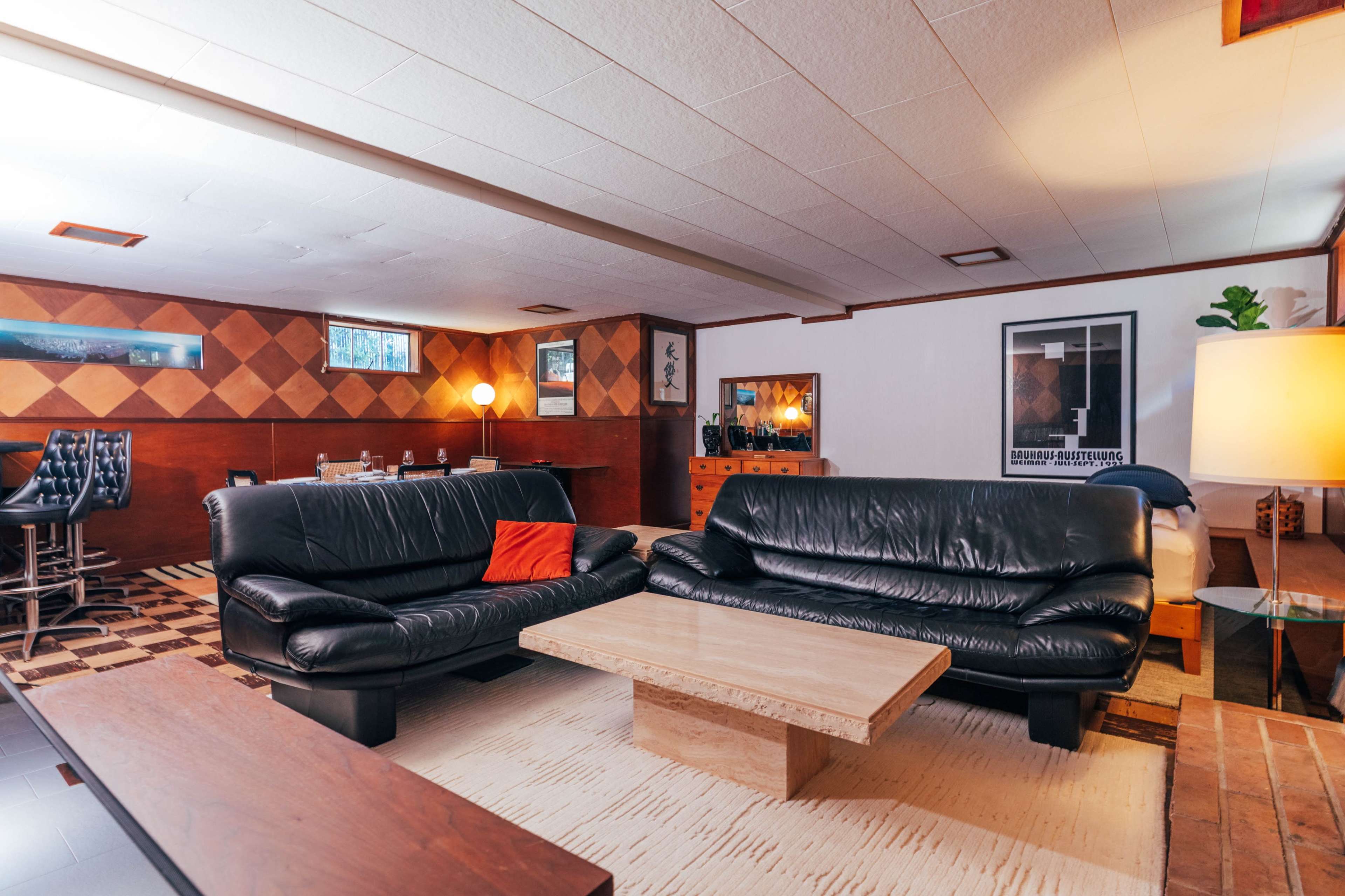 The image shows a modern living room featuring two black leather sofas facing a beige coffee table, with wood-paneled walls and a dining area visible in the background.