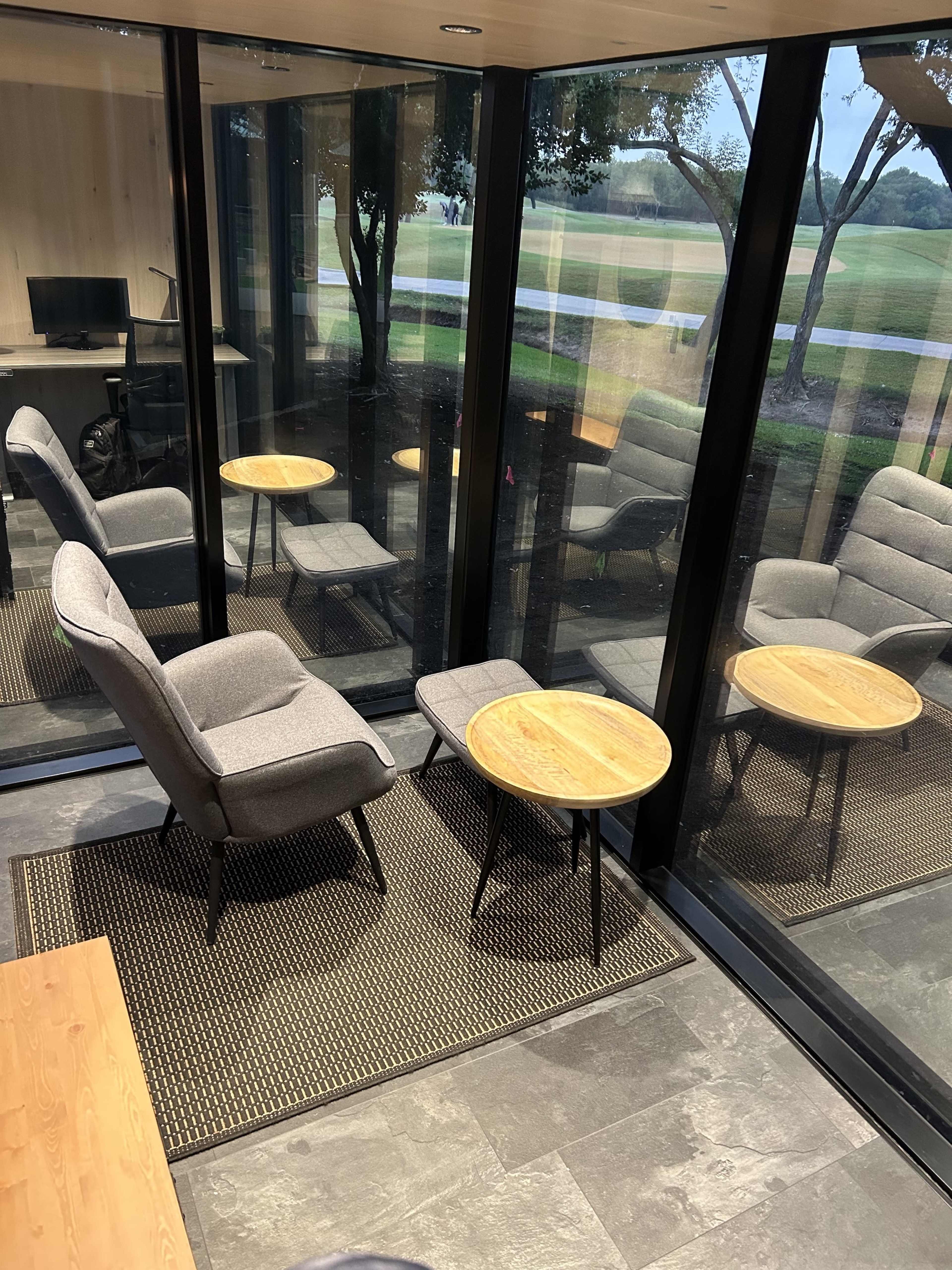 The image shows a secluded sitting area with two armchairs and a small round table, enclosed by glass walls and overlooking a green outdoor space.